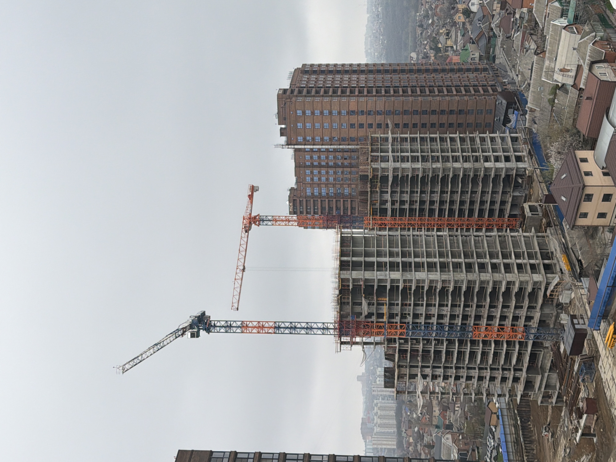 The image shows a construction site in Rostov-on-Don, Russia, with two unfinished high-rise buildings. One building is further along in construction, appearing to have the skeleton completed and the exterior partially enclosed, while the other is a bare concrete frame. Two red and blue tower cranes are visible, presumably aiding in construction. In the background, a fully completed, taller building stands, its facade a brown brick color. The sky is overcast. The background contains cityscape featuring houses and other buildings.