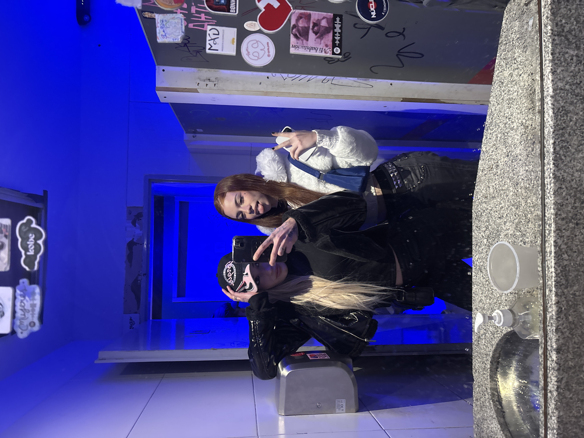 Two women are posing for a selfie in a restroom setting. The restroom has a modern aesthetic, with blue lighting and a gray countertop. The women appear to be young adults or teenagers.
One woman has blonde hair and is wearing a black cap, a black jacket, and black pants. She is holding up her phone to take the picture. The other woman has auburn hair and is wearing a white jacket, a blue bag, and black pants. She has her tongue sticking out in a playful manner.
The background includes a mirror decorated with stickers and writing. To the right of the mirror, there is a sink and a soap dispenser.
The restroom is possibly located in the city of Buenos Aires, Argentina; however, no details in the photo confirm this statement.