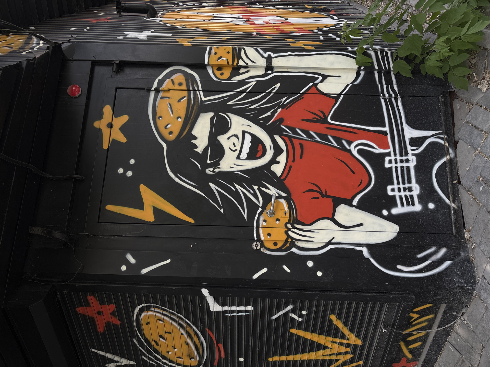 The image depicts a graffiti artwork on the exterior of a building in Novosibirsk, Russia. The graffiti features a stylized cartoon character with long, flowing hair, sunglasses, and an open mouth, seemingly singing or shouting. The character is wearing a red t-shirt and is holding a hamburger above its head with one hand, while another hamburger is held in its outstretched hand. A simplified representation of an electric guitar is painted below the character. The background of the graffiti is black and includes various abstract elements like yellow lightning bolts, red and yellow stars, and white dots.
The building itself appears to be a structure with dark, corrugated metal siding and a dark door, which is part of the graffiti. The door has a silver handle. To the left of the door, a roll-up metal shutter is visible, also adorned with graffiti, including a red star and yellow radiating lines. To the right, the corrugated metal siding continues, with more graffiti elements, including red and yellow patterns.
The ground in front of the building is paved with cobblestones. A small patch of green vegetation is growing at the bottom right corner of the image, partially obscuring the graffiti. Overhead, electrical wires are visible, suggesting an urban setting. The overall scene suggests an artistic and possibly commercial or entertainment-related establishment in an outdoor urban environment.