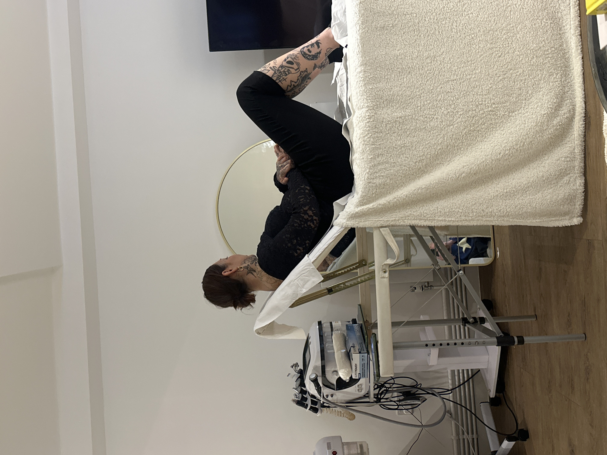 A woman with tattoos is lying on a treatment table in what appears to be a clinic or spa. She's wearing black pants and a black lace top. Her legs are raised. The table is covered with a white towel. There's a machine with wires and various attachments below the table. A round mirror is behind the woman's head. The location is a room with white walls and light-colored wood floors. It's unknown if the image was taken in London, United Kingdom.