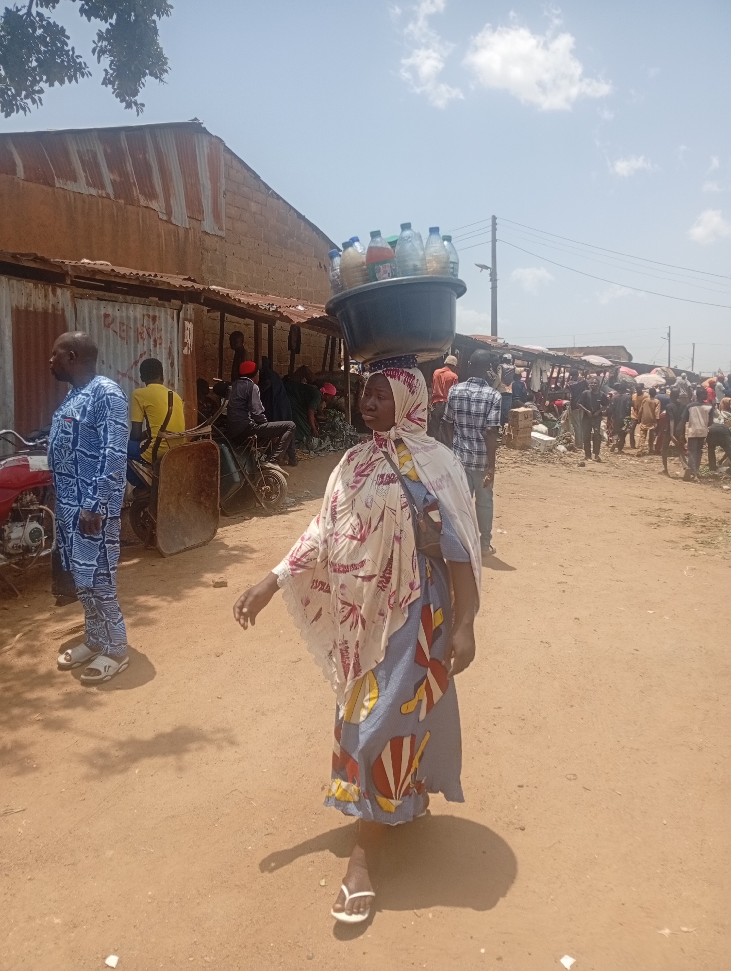 A medium shot shows a woman carrying a container full of water bottles on her head at a crowded outdoor market in an unspecified city, perhaps in Africa. The woman, who is in the foreground, is wearing a long, patterned blue and white dress, a printed headscarf, and white flip-flops. She is facing left with her right arm extended. On her head, she is carrying a dark container filled with approximately ten clear plastic water bottles. To the left of the woman stands a man in a blue patterned shirt and light-colored sandals, facing away from the camera. Behind them are various people and vehicles, including a motorbike and a wheelbarrow. In the background, more people are milling about the market area, which is lined with rudimentary stalls and buildings with rusted metal roofs. The ground is dirt and sand. The sky is partly cloudy.