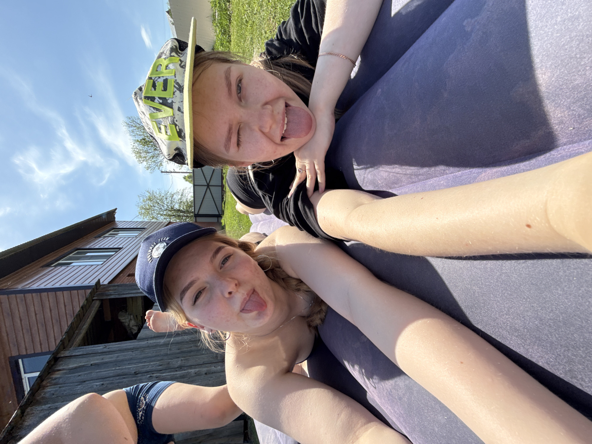 Two young women are posing for a selfie outdoors. One woman is wearing a black shirt and a white, camouflage-patterned cap with yellow lettering. She is lying on a grey or purple surface and has her tongue sticking out while winking. The second woman is wearing a navy blue cap with white lettering and is also sticking her tongue out. Part of a wooden house is visible in the background, along with a blue sky and some greenery. The location appears to be in a rural or semi-rural area. Zhigalki, Russia is mentioned as the location.