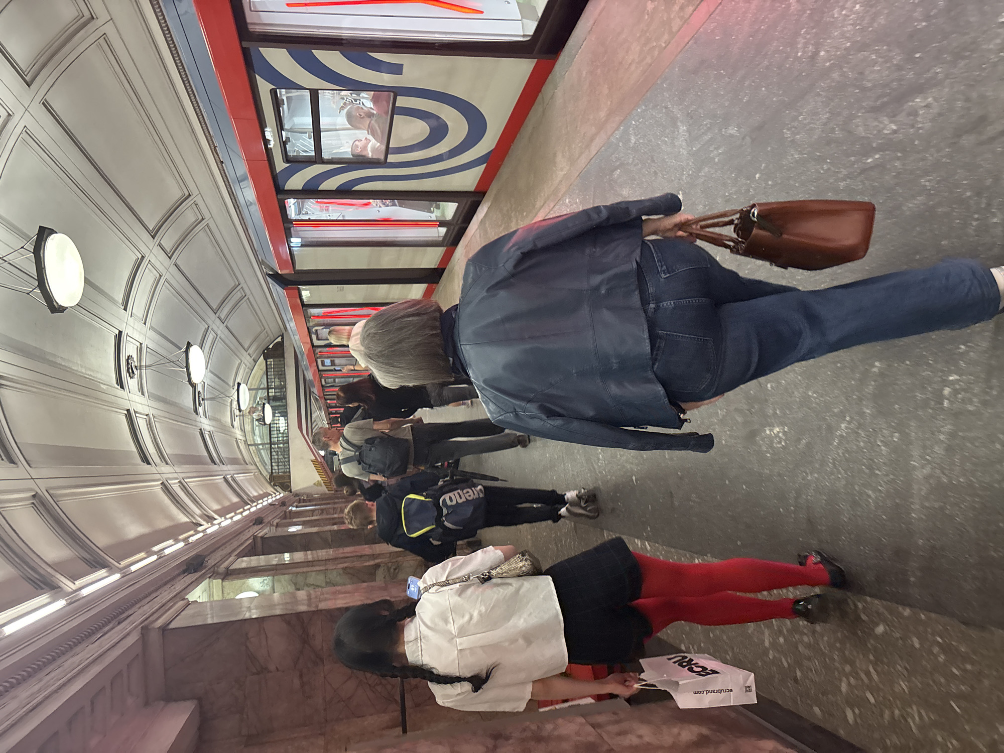 A group of people are walking along a subway platform in Khimki, Russia. The platform is lined with marble columns and a white tiled ceiling with arched panels. A modern train with red and blue markings is stopped at the platform. The people are walking away from the camera, and their faces are not visible. One woman in the foreground is wearing a blue leather jacket and jeans, and carrying a brown handbag. Another woman in the mid-ground is wearing a white shirt, black skirt, and red tights, and carrying a white shopping bag. A young boy in the mid-ground is wearing a backpack. The lighting on the platform is provided by overhead lights and the train's interior lights. The overall scene is one of everyday transit in a public space.