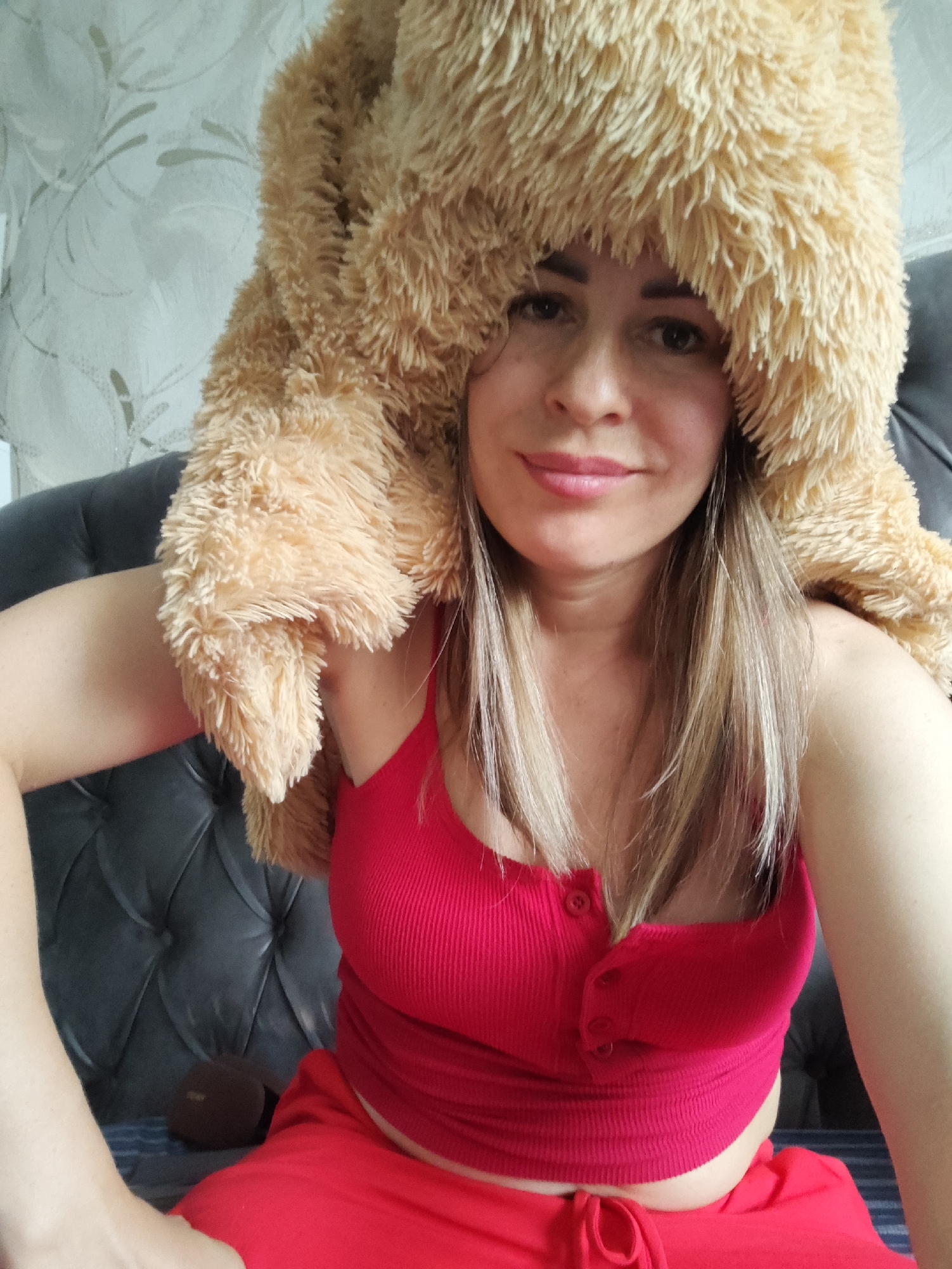 This is a medium shot, selfie-style photograph of a woman sitting in a room. She has fair skin, dark brown hair with blonde highlights, and is wearing a fluffy, light brown, plush hat that covers her head and shoulders. She is also wearing a red tank top with buttons down the front and red shorts. The woman is looking directly at the camera with a slight smile.
The setting appears to be indoors, likely a living room, with a dark leather tufted couch behind her and a patterned wallpaper on the wall. To the left of the woman, there is a dark, oval-shaped object on the couch. The lighting is natural, suggesting it could be daytime.
The woman seems to be in a relaxed and playful mood, indicated by her attire and expression. There is no visible text in the image. The context provided indicates the location is Almaty, Kazakhstan.