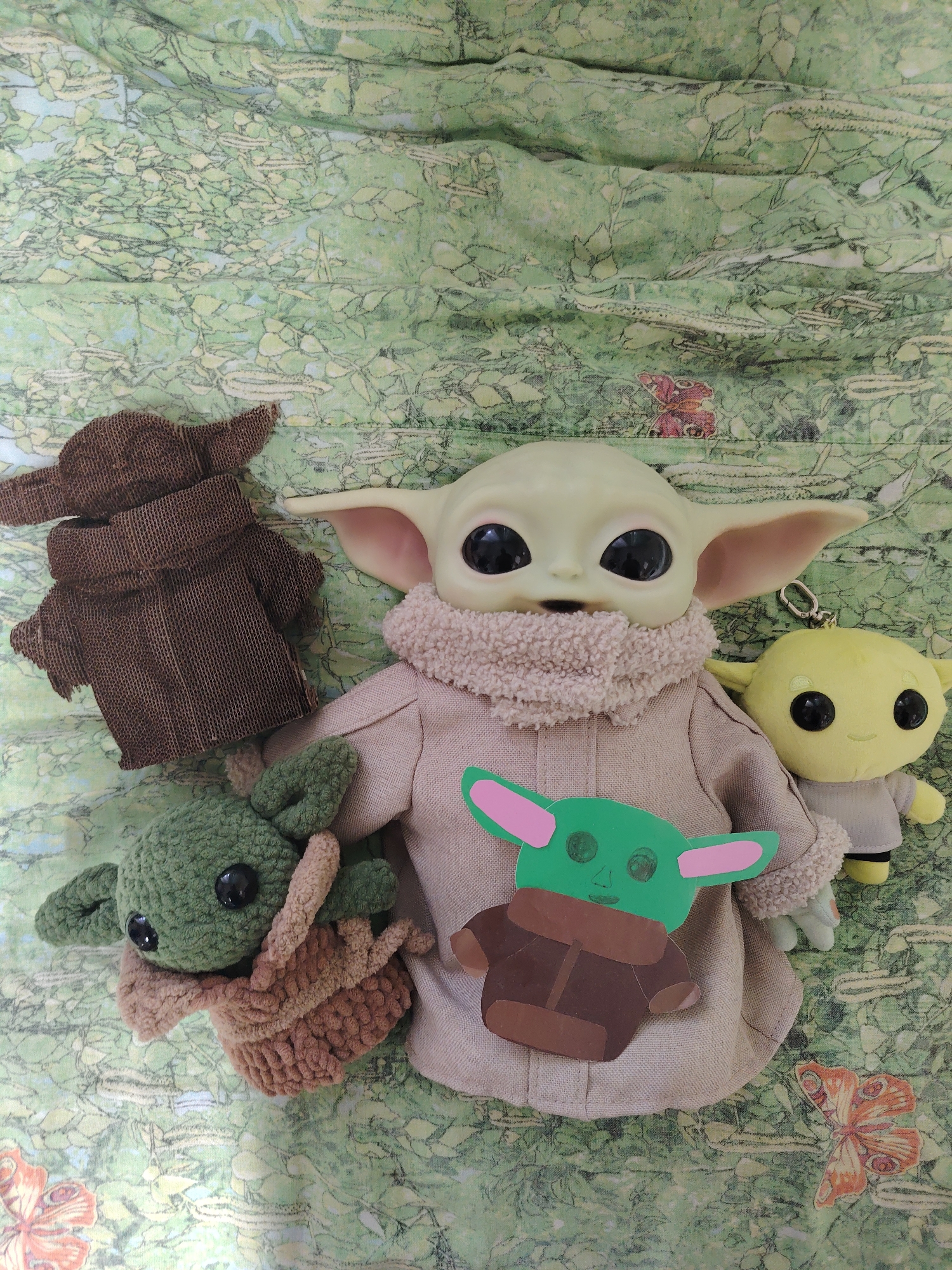 The image shows four Baby Yoda figures on a green patterned surface. The surface seems to be patterned with green leaves and a few butterflies are on the lower edges of the picture. There is a large plastic figure of Baby Yoda in the center, wearing a beige robe with a fluffy collar. A green and brown paper cutout of Baby Yoda is pinned to the larger figure's chest. To the left of the large figure is a brown cardboard cutout figure of Baby Yoda. Below and slightly to the left of the central figure is a knitted or crocheted green Baby Yoda. To the right of the main figure is a small, yellow plush Baby Yoda keychain. No people are present in the image. There is no information to suggest this image was taken in Chaikovsky, Russia.