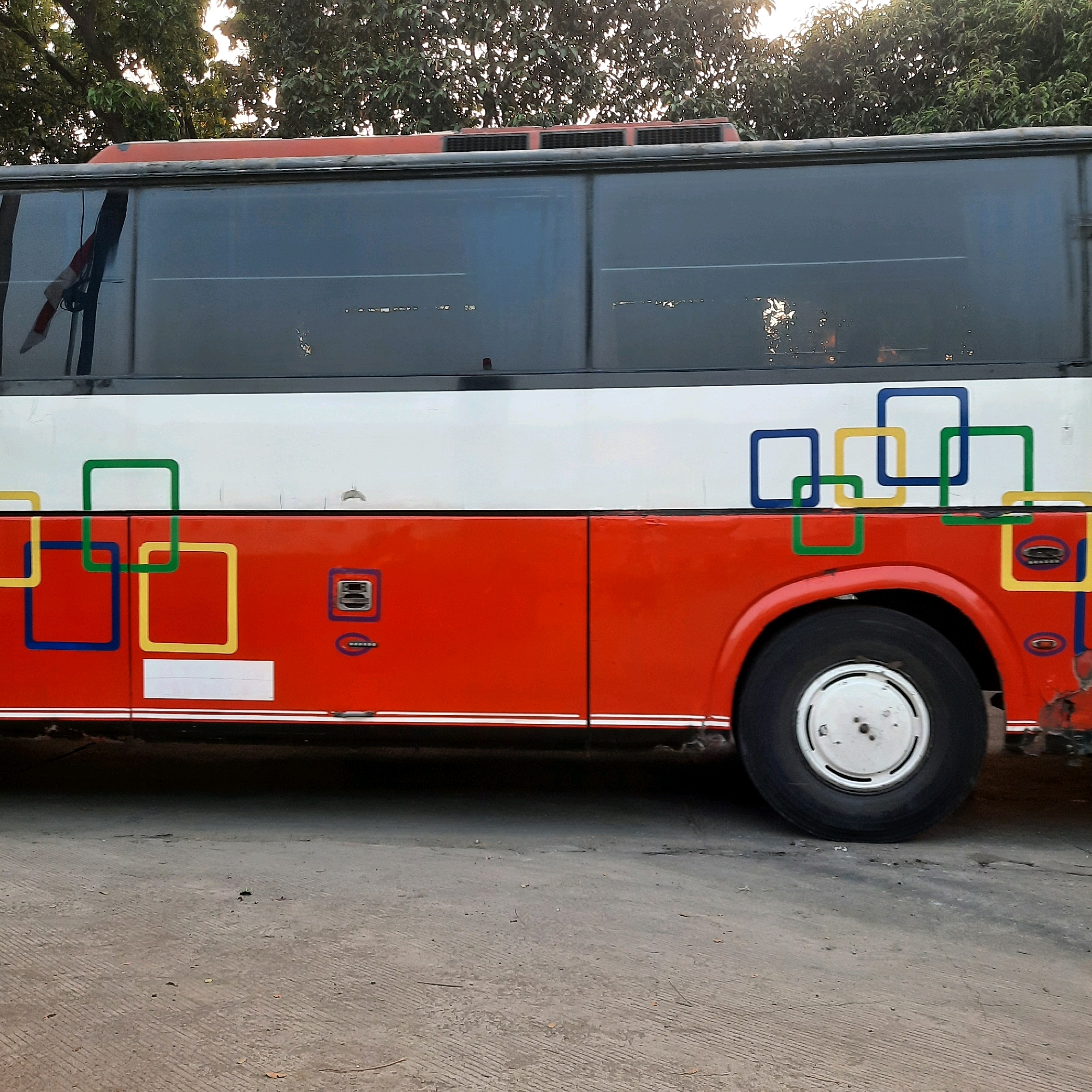 An orange and white bus is parked on a paved surface. The bus has large tinted windows and a decorative pattern of overlapping colored squares on its side. The bus is facing right and the front half of the bus is visible. The foreground is a rough, grey paved surface with some debris. In the background, there are trees with sunlight filtering through the leaves. The bus appears to be in a city environment, possibly East Jakarta, Indonesia. There are no people visible in the image. The bus exhibits some signs of wear and tear, particularly on the lower right side.