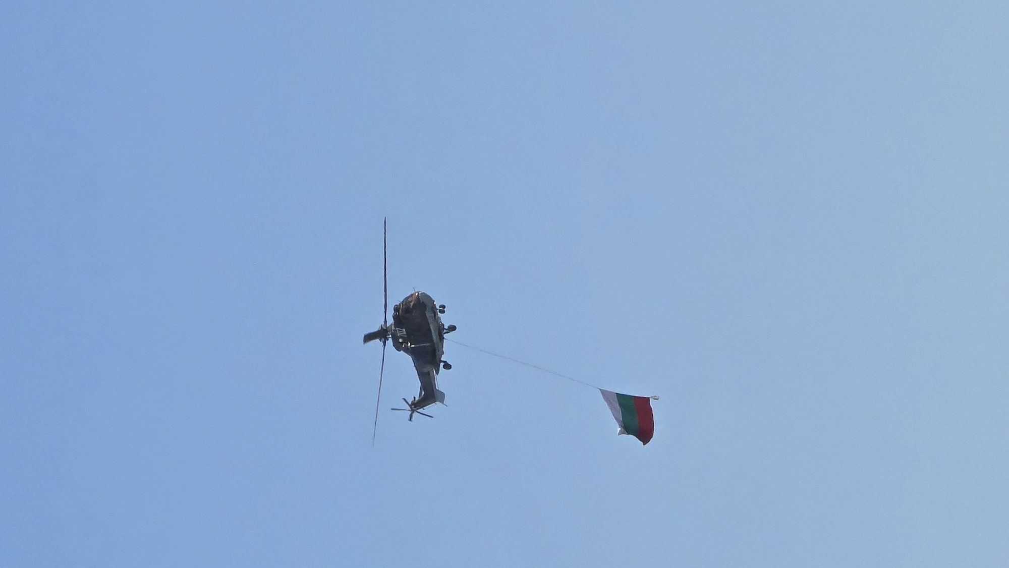 A military helicopter flies in a clear blue sky above Sofia, Bulgaria, carrying the Bulgarian flag. The flag is attached to the helicopter by a rope. The helicopter's rotor blades are spinning.