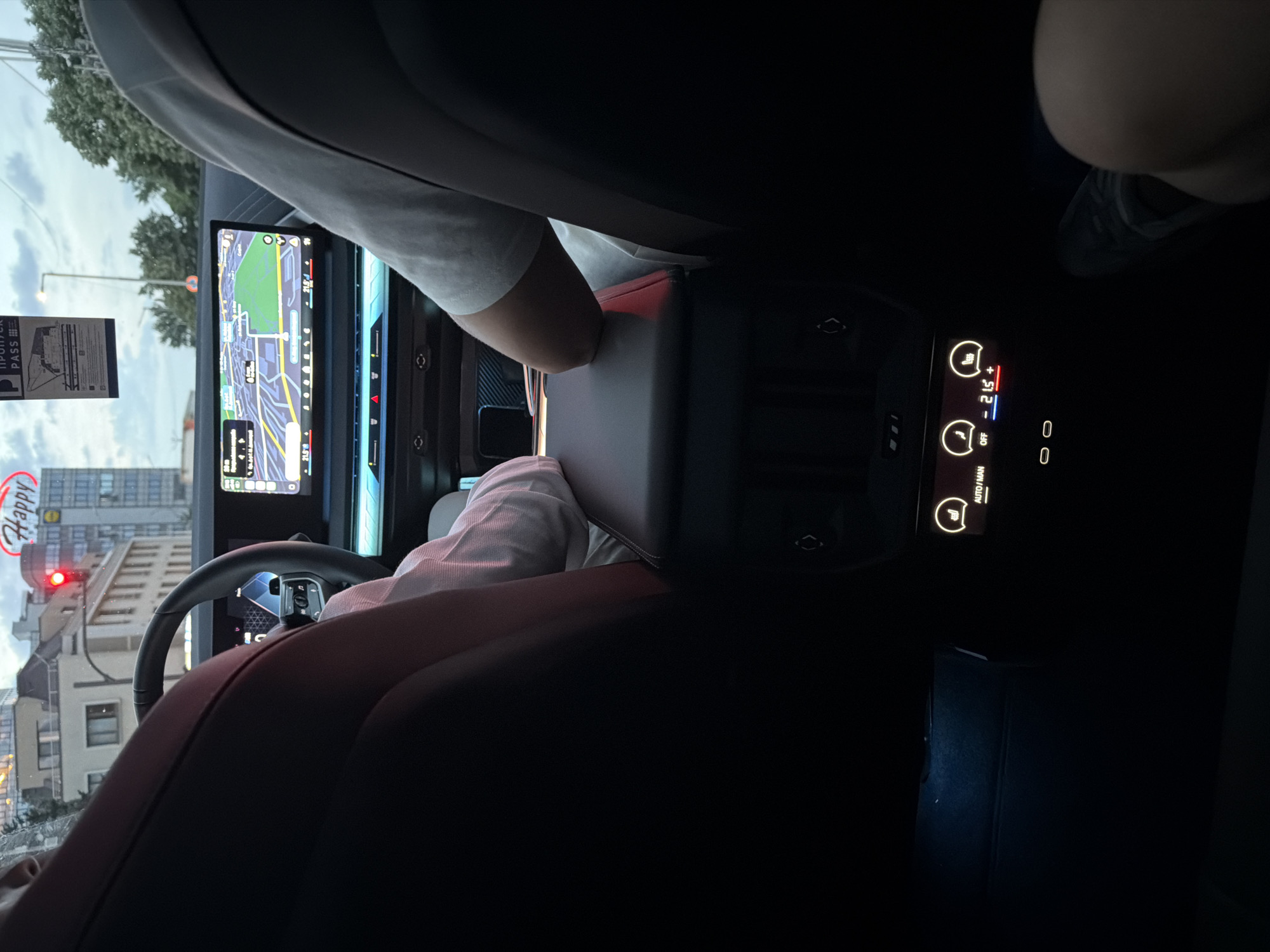 The image provides a detailed view from the rear passenger seat of a modern car, looking towards the front cabin and the external environment in Sofia, Bulgaria.
Inside the vehicle, a driver, clad in a light-colored long-sleeved shirt, is seated on the left, while a front passenger, wearing a light-colored short-sleeved shirt, occupies the right. Both have their arms resting on the shared center console. The car features a sophisticated dashboard with an integrated wide digital display system; the central portion displays a navigation map showing roads and green areas, with a temperature reading of 21.5° Celsius. Below the main screen, a horizontal light-blue ambient light strip highlights a control panel. For rear passengers, a dedicated control unit is visible, featuring illuminated icons for heated seats, a temperature display showing "OFF - 21.5° +", and two USB-C charging ports. A white permit, bearing the Cyrillic inscription "ПРОПУСК PASS !!!", is affixed to the top center of the windshield.
Outside the car, the scene depicts an urban environment under an overcast sky, likely during late afternoon or early evening. The car is stopped at a red traffic light. Multi-story buildings dominate the background, including one prominently displaying a red "Happy" neon sign and a partially visible "Lidl" sign, confirming the location as Sofia, Bulgaria. Trees are visible on the right side of the exterior view. A leg is partially visible in the bottom right corner of the frame, indicating the presence of at least one rear passenger who is likely taking the photograph.