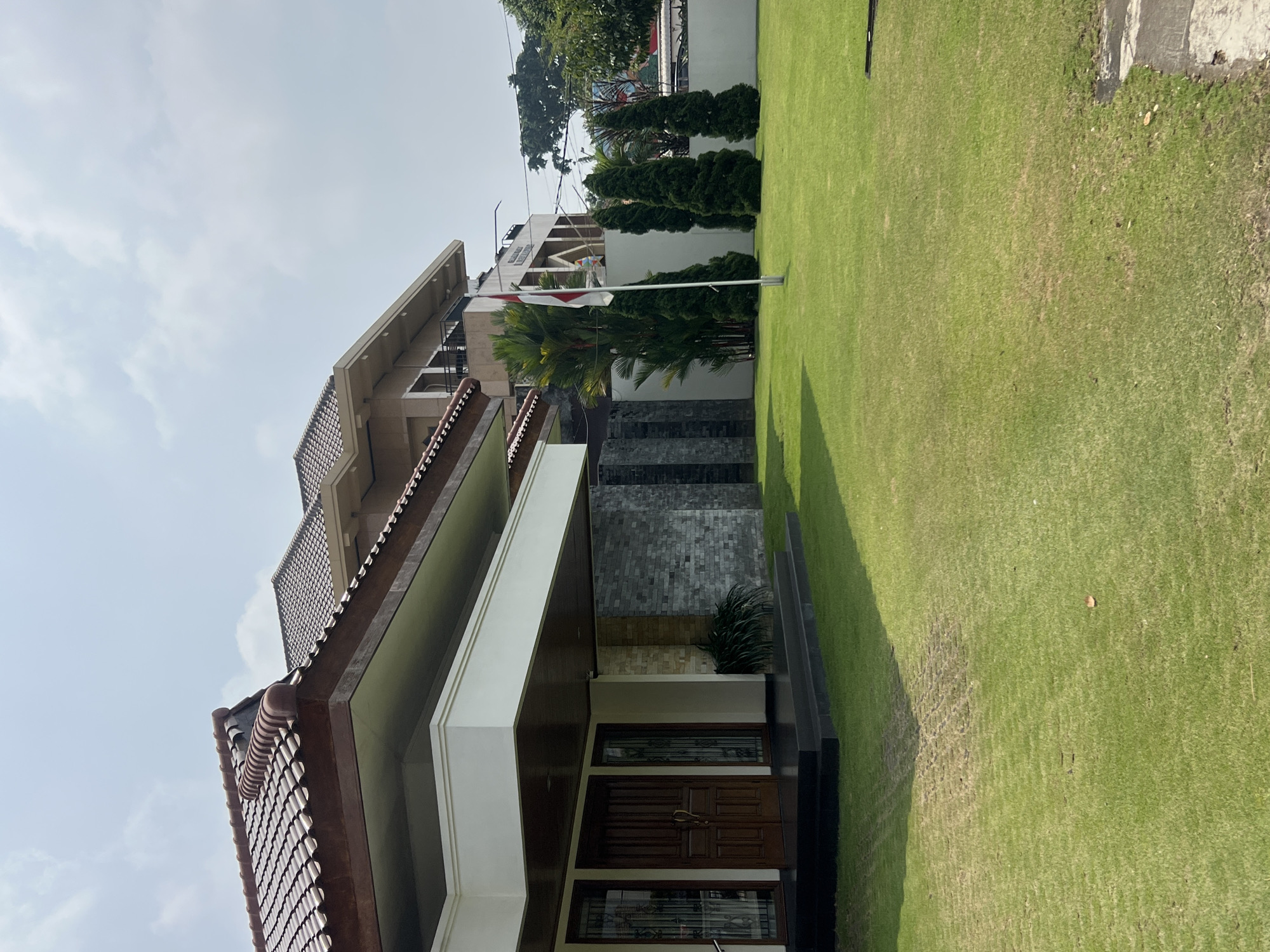 The image shows a large, modern house with a well-maintained lawn in Yogyakarta, Indonesia. The house has a brown tiled roof and white walls with dark brown trim. The entrance features double wooden doors and is shaded by a large overhang. The facade also includes sections of grey stone tiling. The lawn is vibrant green and appears to be freshly mowed. In the background, a flag with red and white stripes, likely the Indonesian flag, flies on a flagpole in front of a second building. Several tall, dark green coniferous trees are planted in a line on the property, and other tropical foliage is visible. The sky is partly cloudy. The overall scene conveys a sense of affluence and order.