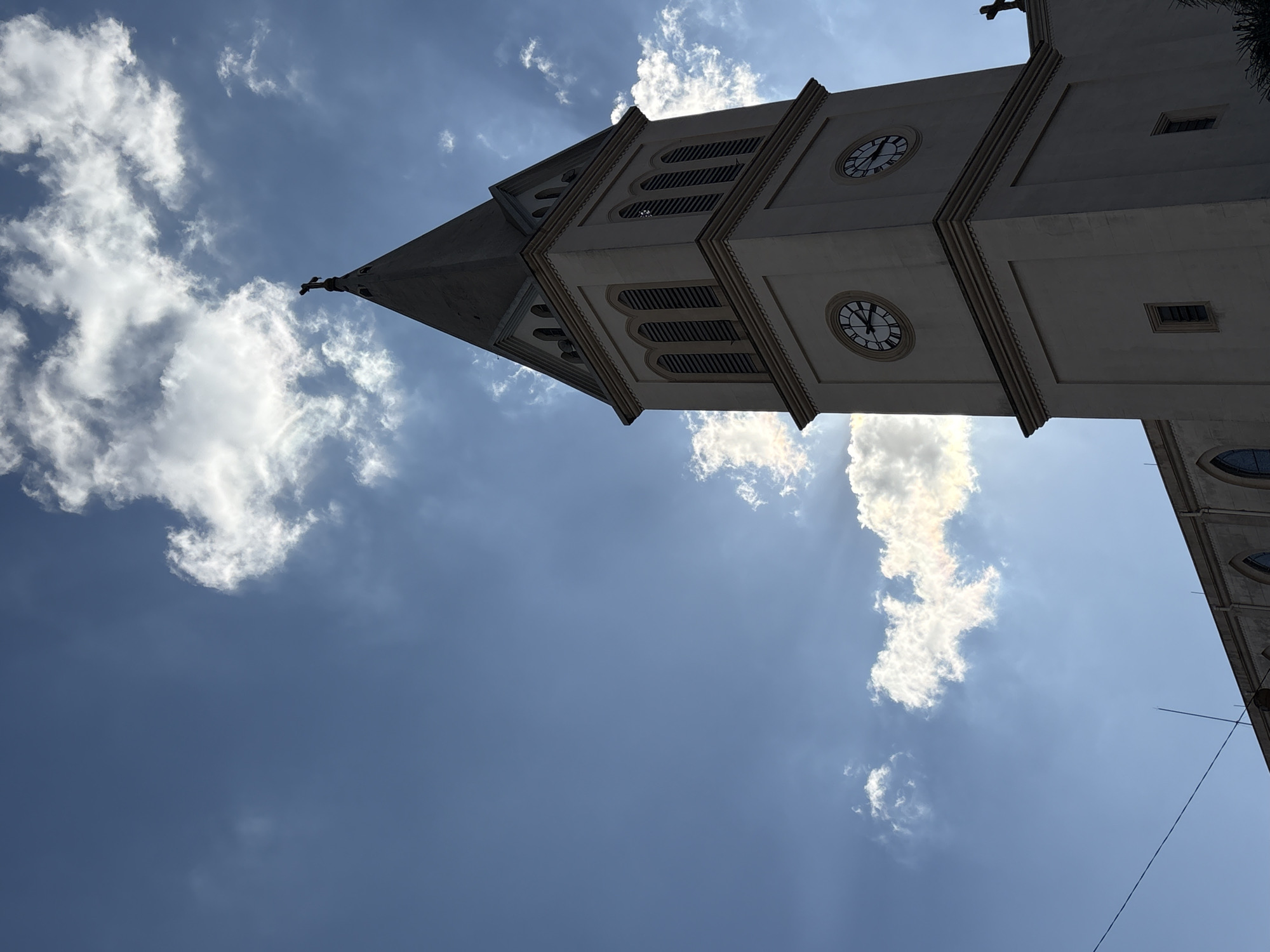 This is a low-angle, silhouetted photograph of the tower of a church against a bright blue sky with scattered clouds. The church tower is tall and slender, with a pointed steeple topped by a cross. The tower has at least two clock faces, one on each visible side, showing roman numerals. The sun is behind the tower, creating a silhouette effect and highlighting the clouds with rays of light. Part of the church's roofline is visible in the foreground, also silhouetted. The image appears to be taken in an urban setting, as suggested by the power line visible in the lower left of the frame. The church tower has architectural details typical of European Gothic or Neoclassical styles, with arched window openings and decorative cornices. The overall lighting suggests a bright, sunny day.