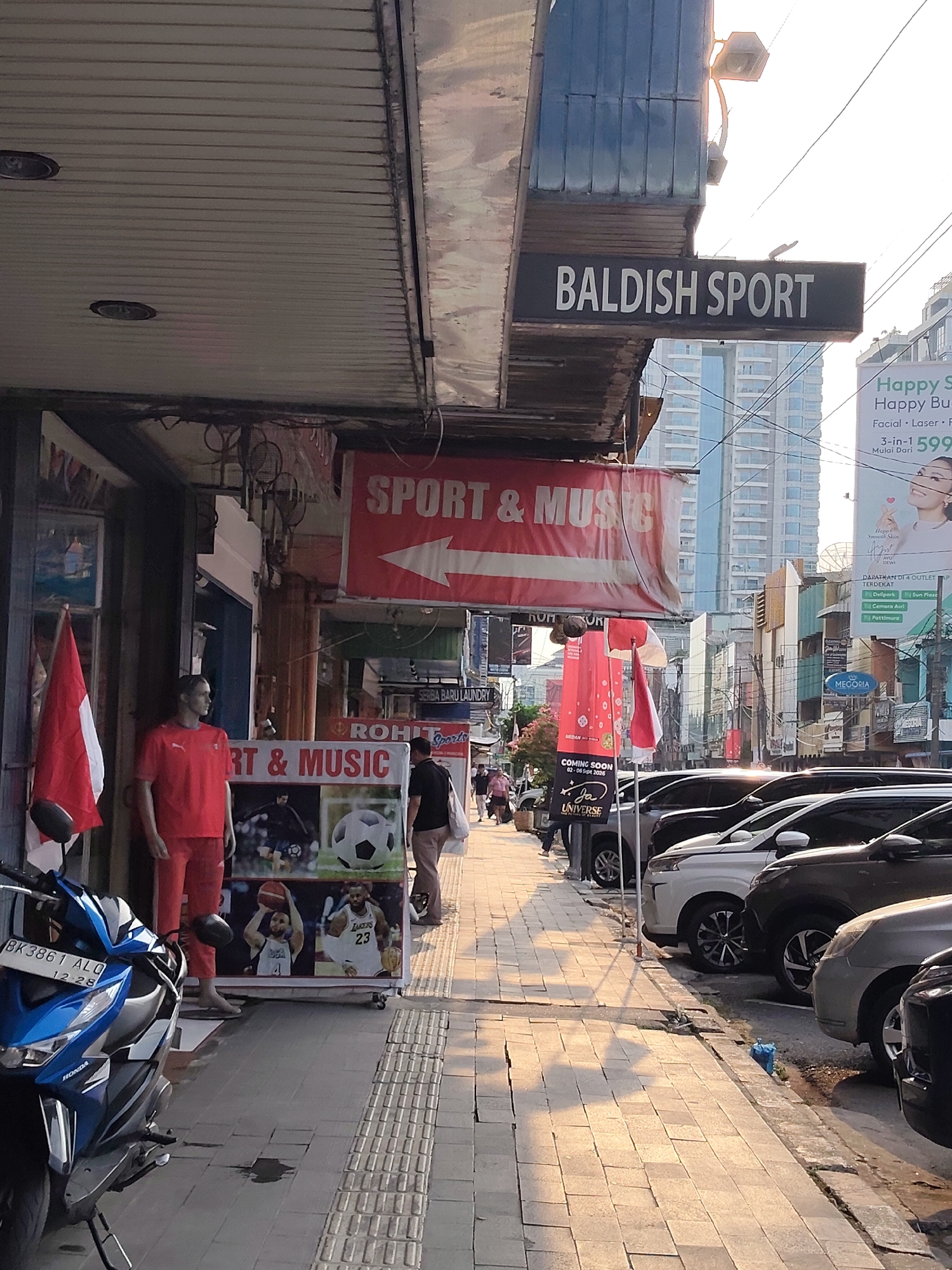 This is a street-level photograph taken in the city of Medan, Indonesia. The scene depicts a sidewalk in front of several storefronts. A prominent sign above reads "BALDISH SPORT". To the left, a mannequin wearing a red sportswear outfit stands near a display featuring images of a soccer ball and athletes. A blue and white Honda motorcycle is parked on the left side of the sidewalk. The sidewalk is paved with square tiles and has tactile paving strips. Cars are parked along the street on the right side of the image. The background shows buildings of varying heights and a bright sky, suggesting it is daytime.
