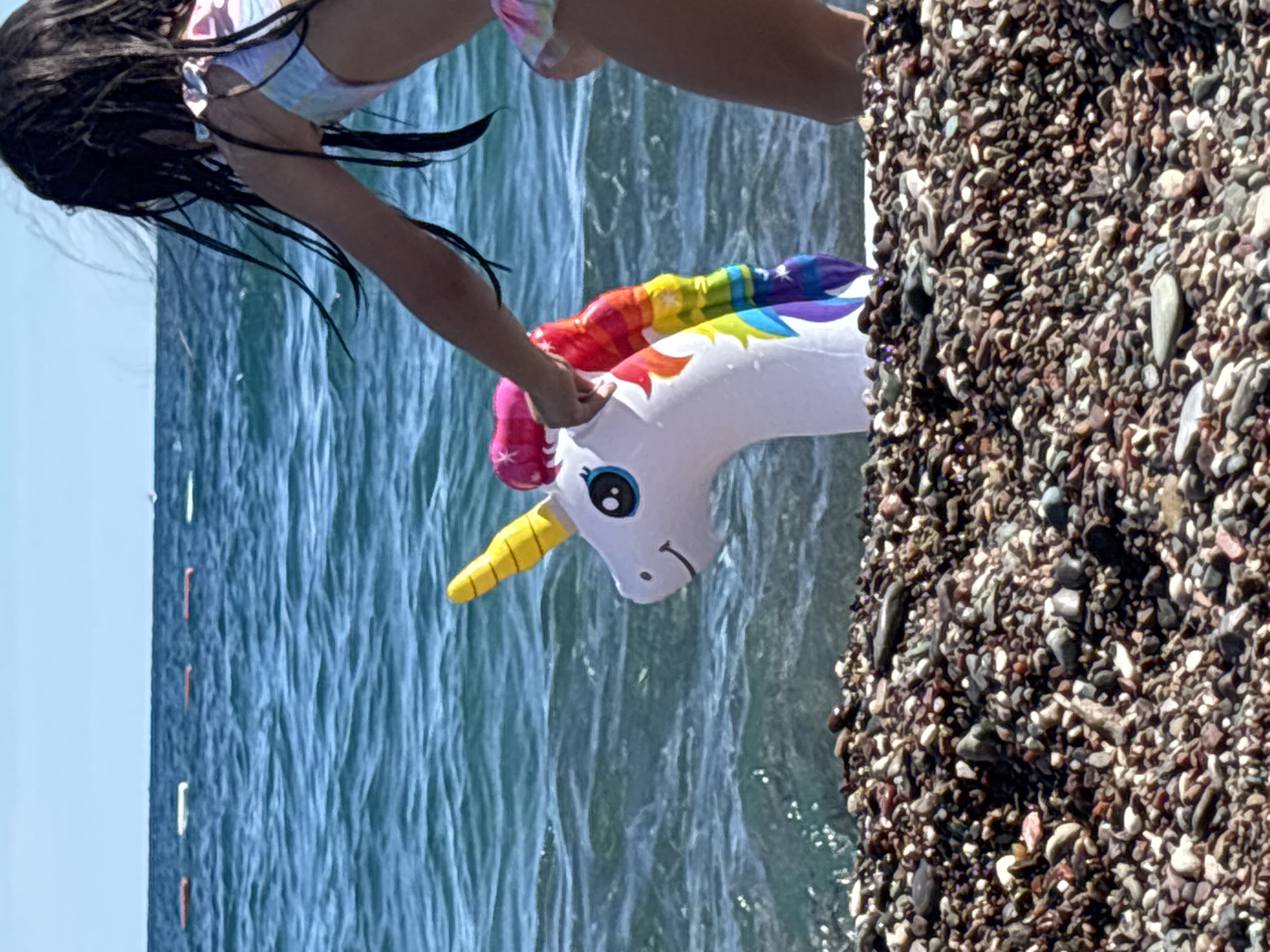 A person, likely a child given the size and swimwear, is partially visible at the top of the frame. They have long, dark, wet hair and are wearing a bikini with a colorful, tie-dye pattern. Their arms are extended towards an inflatable unicorn toy floating in the water. The unicorn is white with a rainbow-colored mane and a yellow horn. It has black cartoon-like eyes and a simple black mouth.
The scene is a beach in Boreti, Montenegro. The water is a clear, deep blue with visible ripples and waves, suggesting it is daytime. The sun is bright, casting a slight glare on the water's surface. To the right of the image, there is a prominent bank of dark, small, smooth pebbles, characteristic of a pebble beach. In the background, the vast expanse of the sea stretches out, meeting a clear blue sky. There are also faint vertical lines in the water to the left, possibly buoys or markers. The overall mood is one of leisure and summer enjoyment. No visible text is present in the image.