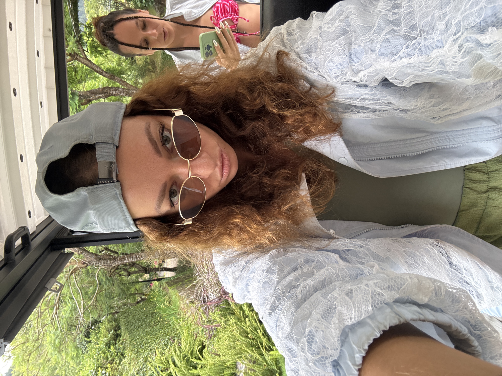 This media file captures a selfie-style shot of two women seated in what appears to be an open-sided vehicle, with a vibrant natural landscape in the background. The image is rotated 90 degrees clockwise.
The woman in the foreground has reddish-brown curly hair and is wearing a light blue baseball cap, with the brand "Jordan" visible on its metallic buckle. She also wears gold-framed sunglasses and a light blue jacket adorned with a delicate white lace overlay, over a green top. She looks directly at the camera with a neutral expression.
Behind her, partially visible, is another woman with dark hair styled in braids. She is wearing a pink and white patterned bikini top under a light-colored, possibly white, top or cover-up. She is holding a light green smartphone with long, white-tipped fingernails, and appears to be looking down at its screen.
The setting is outdoors during the daytime, with bright, natural lighting. The backdrop is a lush, green landscape filled with trees and dense foliage, consistent with the mountainous and forested environment of Púng Luông, Vietnam. The vehicle's interior shows white paneling and black framing, suggesting an open-air mode of transport like a jeep or golf cart. No specific activity beyond posing for a picture and looking at a phone is evident.