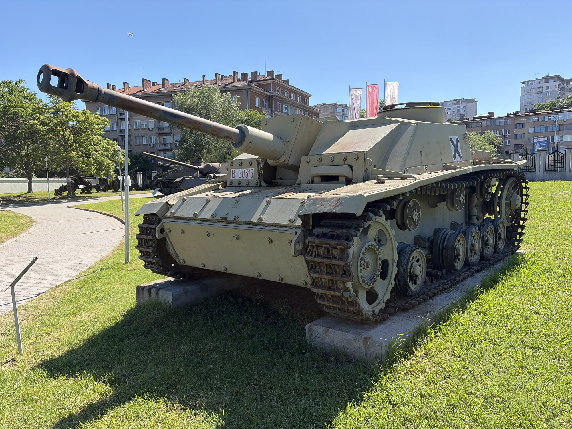 An exterior, eye-level, medium shot captures a German StuG III assault gun displayed outdoors on a sunny day in Sofia, Bulgaria. The vehicle, painted in a dull green camouflage, is positioned on a concrete slab on a grassy lawn. The StuG III's long barrel points to the left, its muzzle brake clearly visible. A license plate reading "B. 60516" is affixed to the front of the vehicle. A black "X" symbol is painted on the side of the superstructure. In the background, several multi-story buildings with red-tiled roofs and a lighter-colored modern building can be seen. Flags, some red and white, are planted in front of one of the buildings. Trees with green foliage dot the landscape. A paved walkway curves into the left side of the frame, with a small metal signpost near its edge. A light-colored airplane is visible high in the bright blue sky. The scene appears to be part of a museum or memorial site, possibly the National Museum of Military History in Sofia, given the presence of historical military vehicles displayed outdoors.
