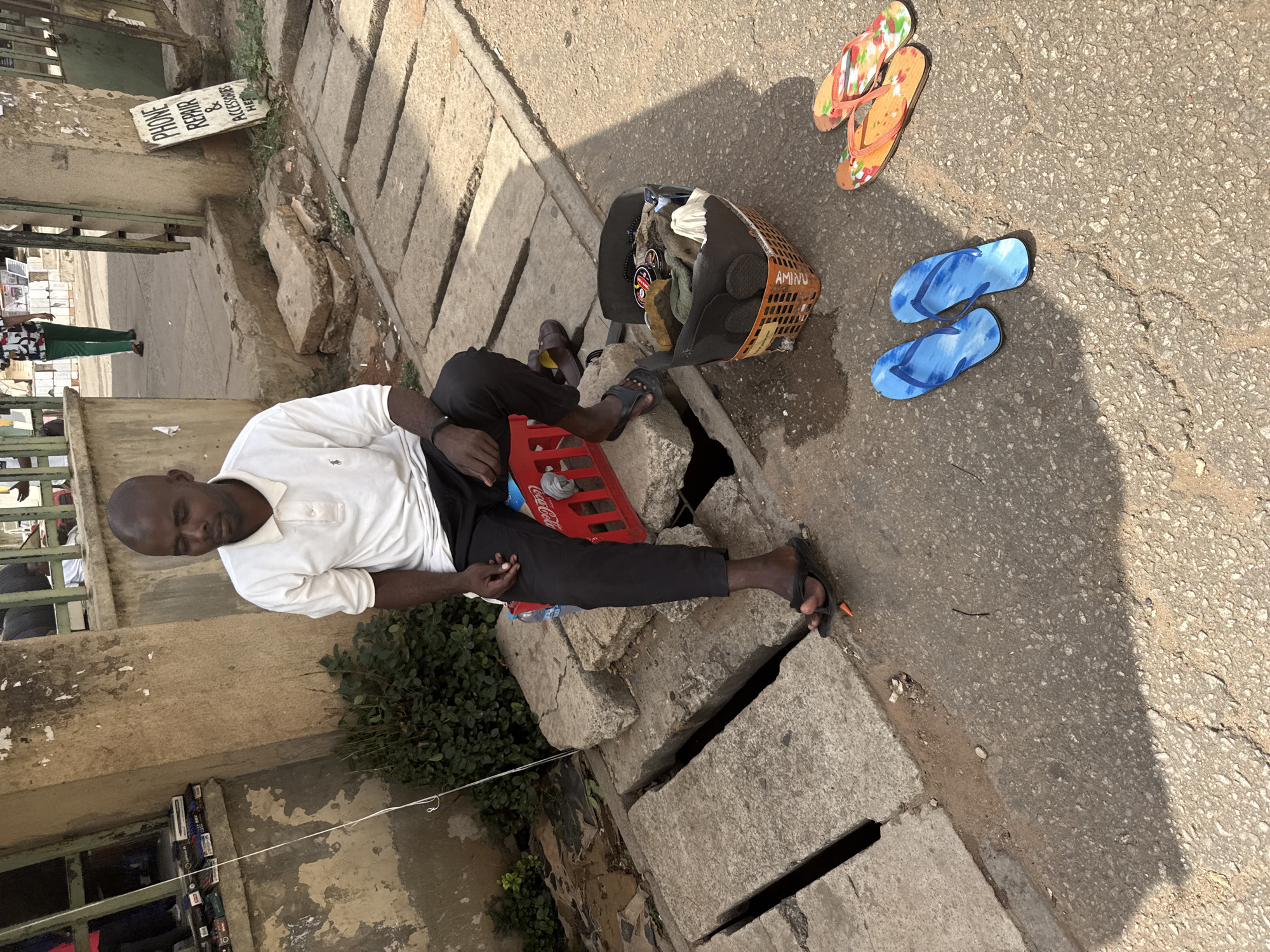 A man is sitting on a concrete barrier next to a drainage ditch in Abuja, Nigeria. He is wearing a white polo shirt and black pants. He is using a red Coca-Cola crate as a seat. Next to him is a plastic basket filled with various items. A pair of blue sandals and a pair of orange and floral sandals are lying on the paved ground. In the background, there's a sign that says "Phone Repair & Accessories Here." The scene appears to be a street-side setting.