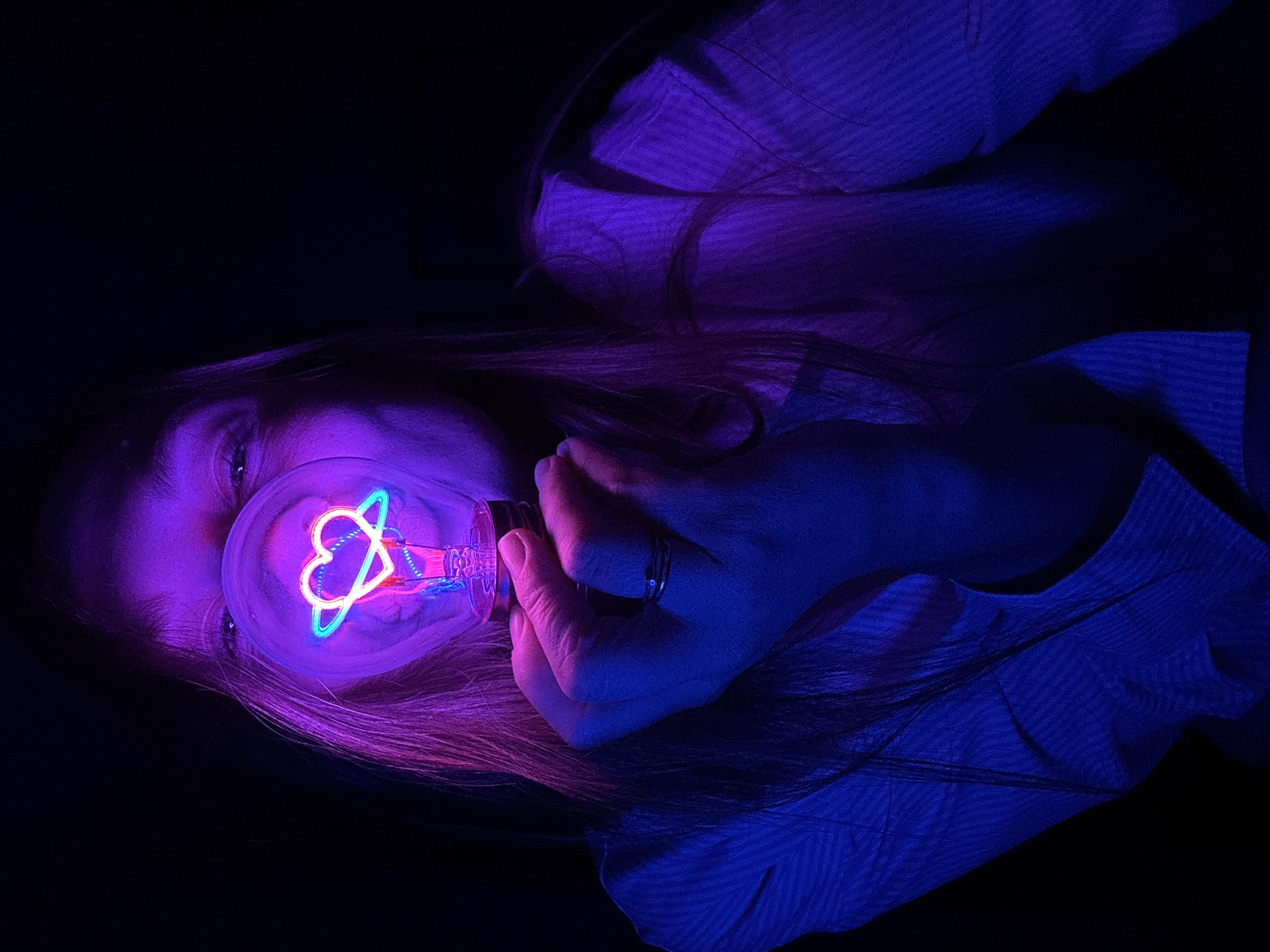 A woman is lying down with a light bulb held up to her face. The light bulb contains a neon heart and a blue ring around it. The scene is lit with purple and blue light. The woman is wearing a striped shirt and a ring on her finger. It is unlikely to determine the specific location in Budva, Montenegro without additional context or information.