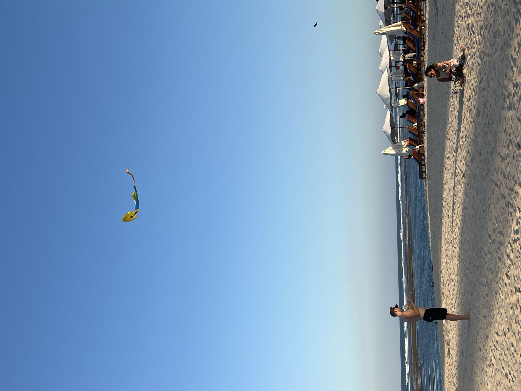 A man stands on a sandy beach in Kuta Selatan, Indonesia, holding a kite string as a yellow kite flies in the clear blue sky. He is shirtless, wearing black shorts, and a black baseball cap. The ocean is visible behind him with waves breaking on a reef. In the background, beach chairs and umbrellas are lined up. A woman and child are sitting on the sand in the foreground, and another bird is visible flying in the distance.