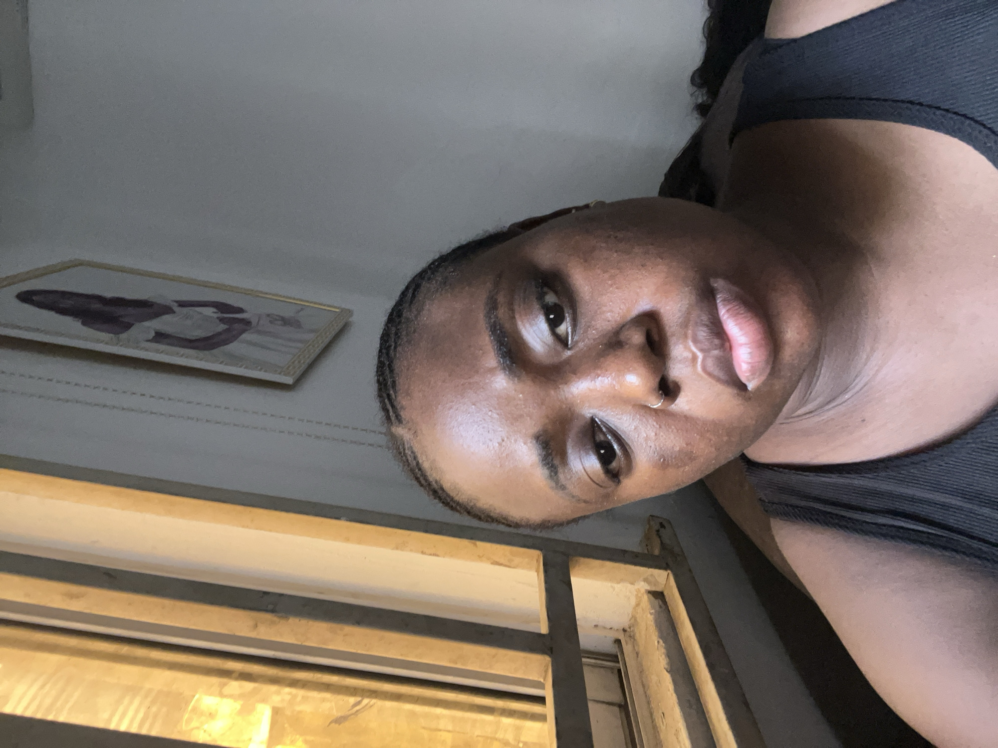 A medium shot of a Black woman is angled towards the top of the frame. She has dark skin, dark eyes, and full lips, and she wears a nose ring. Her hair is styled with braids along her hairline. She is wearing a dark, possibly black, tank top.
The environment includes a wall painted a light color, a framed artwork hanging on the wall, and a window with metal bars. The lighting suggests an indoor environment, possibly a room in a house or apartment. There is insufficient detail to definitively identify the location as Abuja, Nigeria, as this would require specific landmarks or cultural identifiers not present in the image.