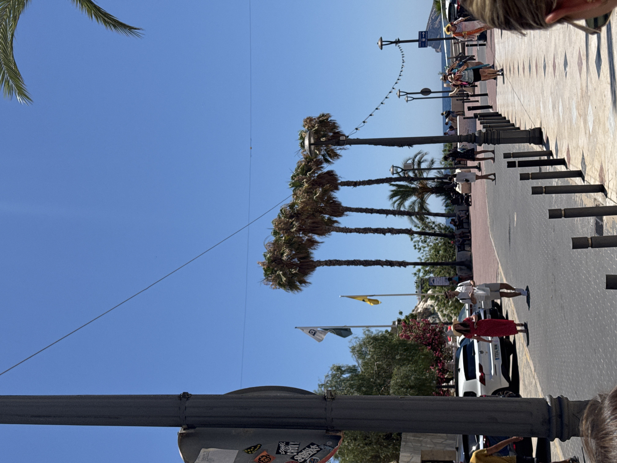 The image shows a sunny day with a clear blue sky and a few wispy clouds. The scene is a paved walkway lined with lampposts and palm trees, suggesting a coastal or resort town. Several people are walking along the walkway, some casually strolling, others appearing to be tourists. In the background, there is a road with parked cars. One white car is prominently visible, with a license plate that reads "7234 NMW". There are also flags visible, one yellow and one with a white emblem and a green stripe, fluttering in the breeze. The overall atmosphere appears relaxed and leisurely, indicative of a holiday or vacation setting. The time of day is likely midday or early afternoon, given the bright sunlight and the long shadows cast by the people and objects. The presence of palm trees and the clear blue sky are strong indicators of a warm climate, consistent with a location like Benidorm, Spain.