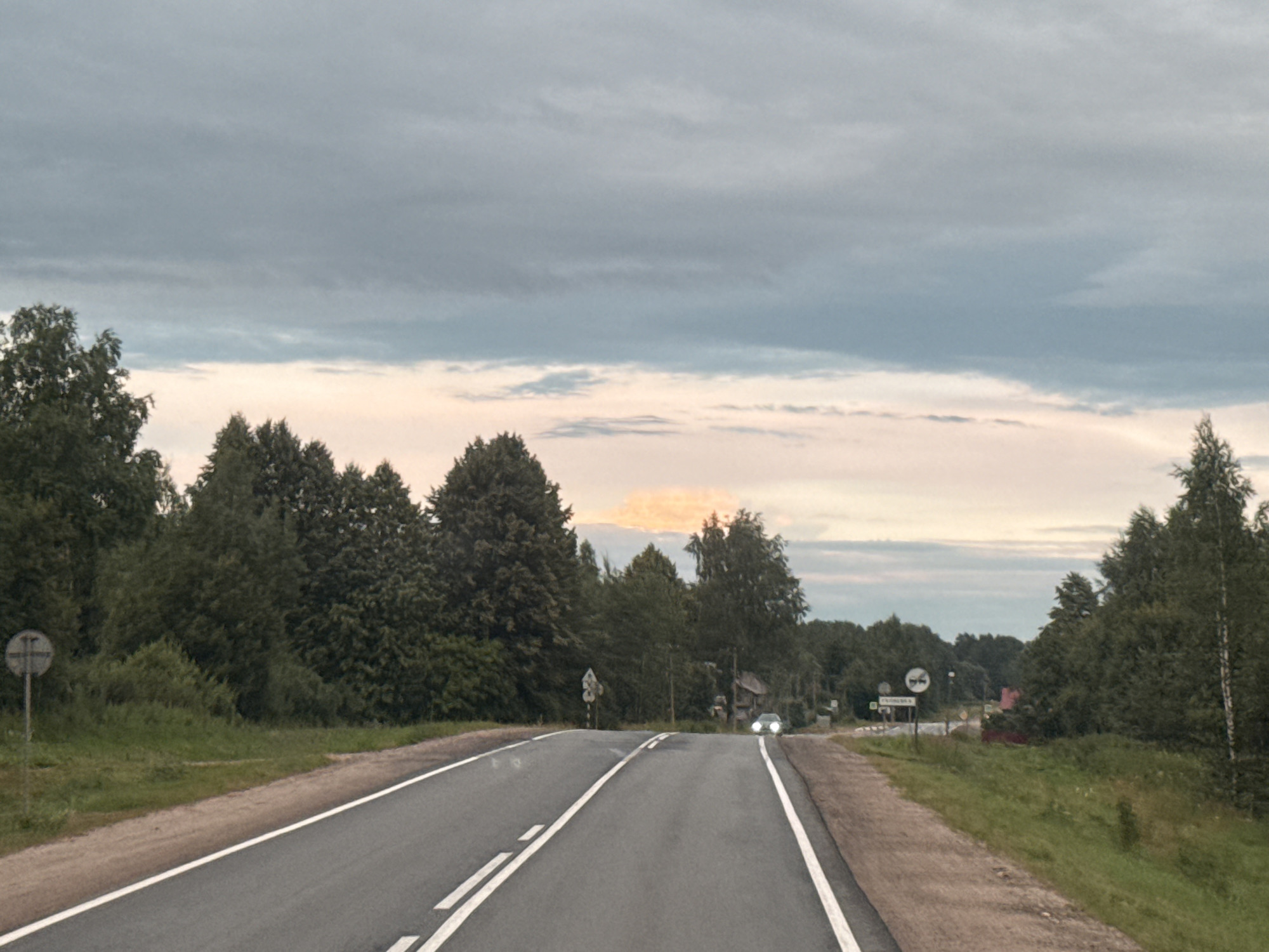 The image shows a two-lane asphalt road with white dashed lines in the center and solid white lines on the edges. The road curves slightly to the right and ascends. On the left side of the road, there is a grassy verge and a dense line of trees. A circular sign with a cross symbol is visible on a pole on the left verge. On the right side of the road, there is also a grassy verge and trees. In the distance, there are more trees, some houses, and a red building. A car with its headlights on is approaching from the distance on the right lane. The sky is overcast with clouds, but there are hints of pink and orange light, suggesting either sunrise or sunset. The scene is rural, and the presence of a road sign with Cyrillic lettering and the general landscape suggest it is located in Russia, specifically in the vicinity of Гуляевка. There are no people visible in the image.
