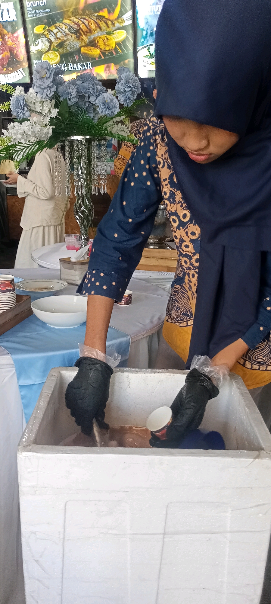 The image captures a close-up of a person wearing a dark blue hijab and a patterned blue and beige top, preparing food or drinks from a white styrofoam cooler. The person is wearing black gloves with clear plastic disposable gloves over them. One hand is submerged in the cooler, while the other holds a small white paper cup. The cooler appears to contain a brownish liquid and possibly ice. In the background, there are posters of food, a floral arrangement with blue and white flowers, and a woman in a light-colored outfit. The scene is set on a table covered with a white tablecloth, with a blue runner. This setup suggests a food service or catering event.
