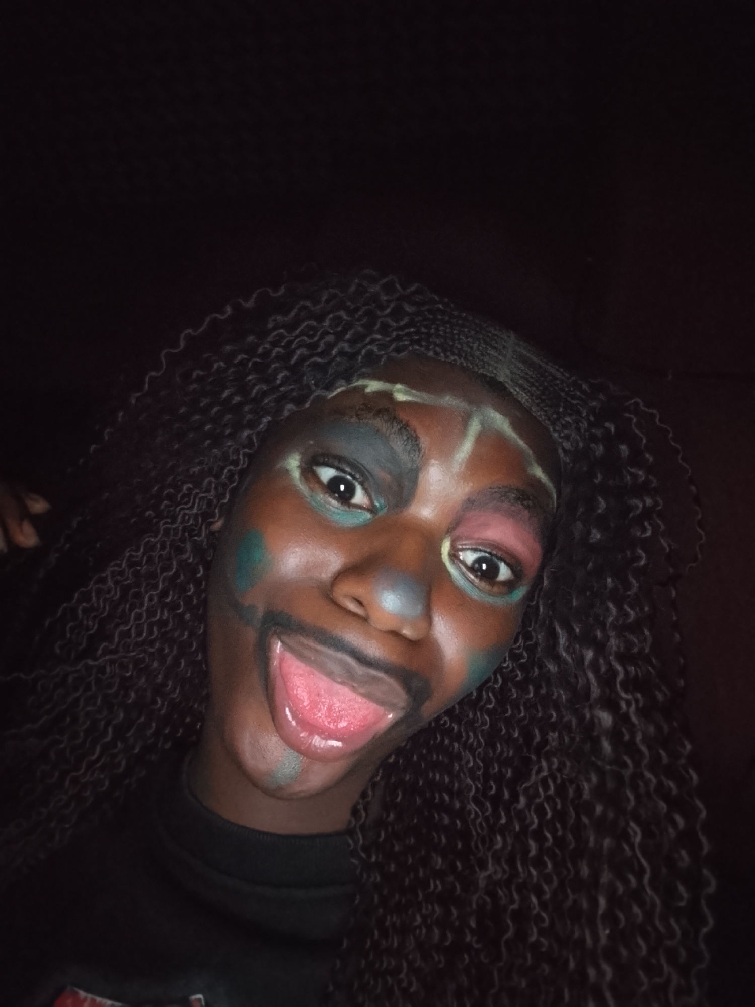 A close-up portrait features a person with dark skin and long, dark braided hair, filling the lower portion of the frame against a completely dark background. The individual is looking directly at the camera with wide, expressive eyes, and their mouth is open with their tongue sticking out playfully.
Their face is adorned with colorful and artistic makeup. The area around their left eye (viewer's left) is covered in a dark blue or black shade, with a bright teal or light green smudge below it. Around their right eye (viewer's right), there's a distinct splash of pink or reddish makeup. Light green or yellowish lines are painted on their forehead, extending upwards and across the brow bone, suggesting abstract patterns or contours. A dark blue-grey smudge is visible on the bridge of their nose, and a lighter grey or green stripe runs down their chin from below the lower lip. The person's lips are a natural pink, and their tongue is a vibrant red.
The lighting is dim, highlighting the face and the colorful makeup against the dark surroundings. The person's expression is one of playful amusement or mischief. They are wearing a dark-colored top, possibly a t-shirt, which is only partially visible at the bottom of the frame. No specific location cues or text are visible in the image.