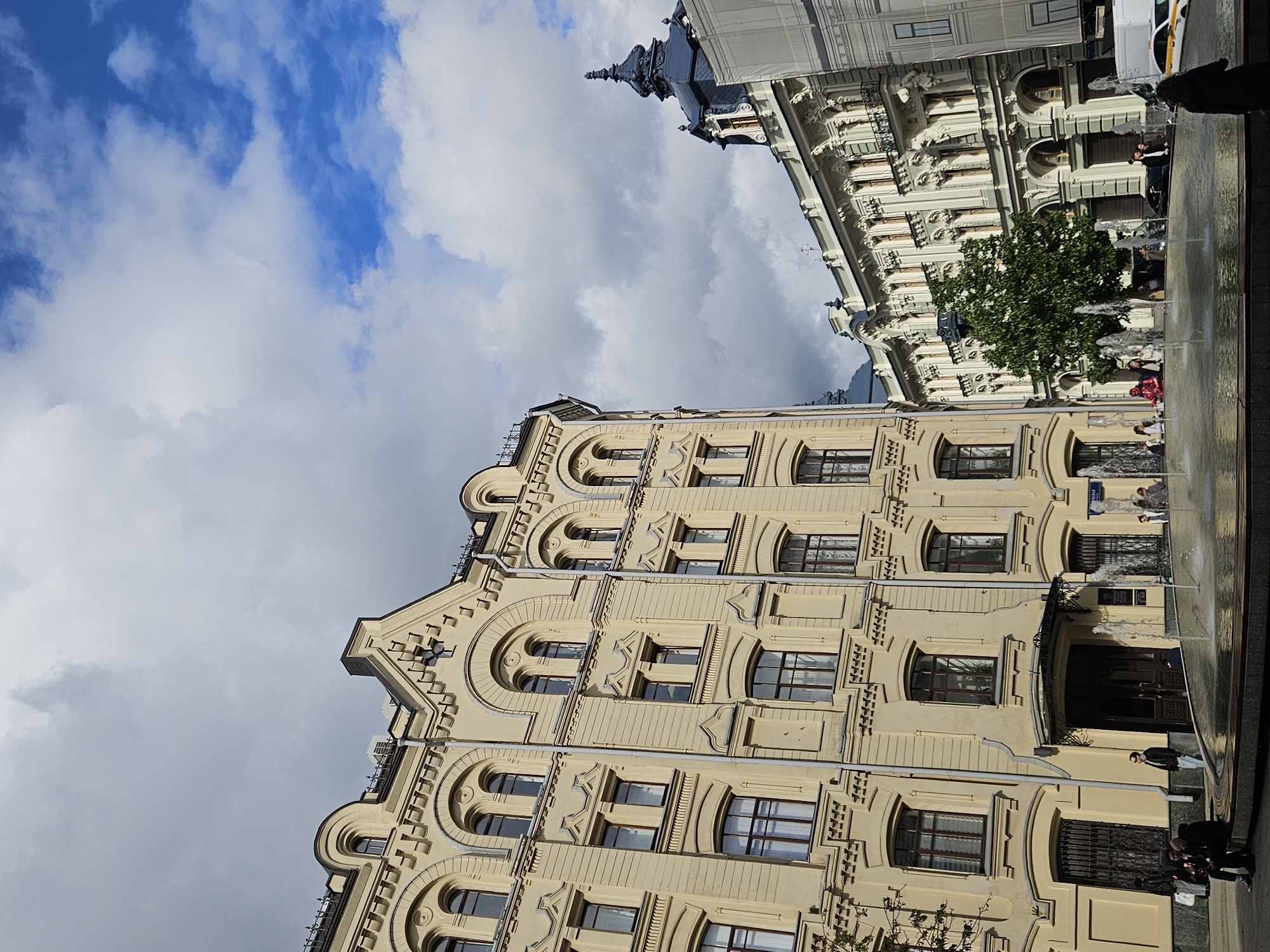 The image shows a street in Moscow, Russia. Two buildings are visible, both with ornate facades. The building on the left is beige and has multiple stories with arched windows. The building on the right is lighter in color with intricate decorative elements. A fountain with multiple jets of water is located in the foreground. Several people are visible walking along the street and interacting near the fountain. The sky is partly cloudy with patches of blue.