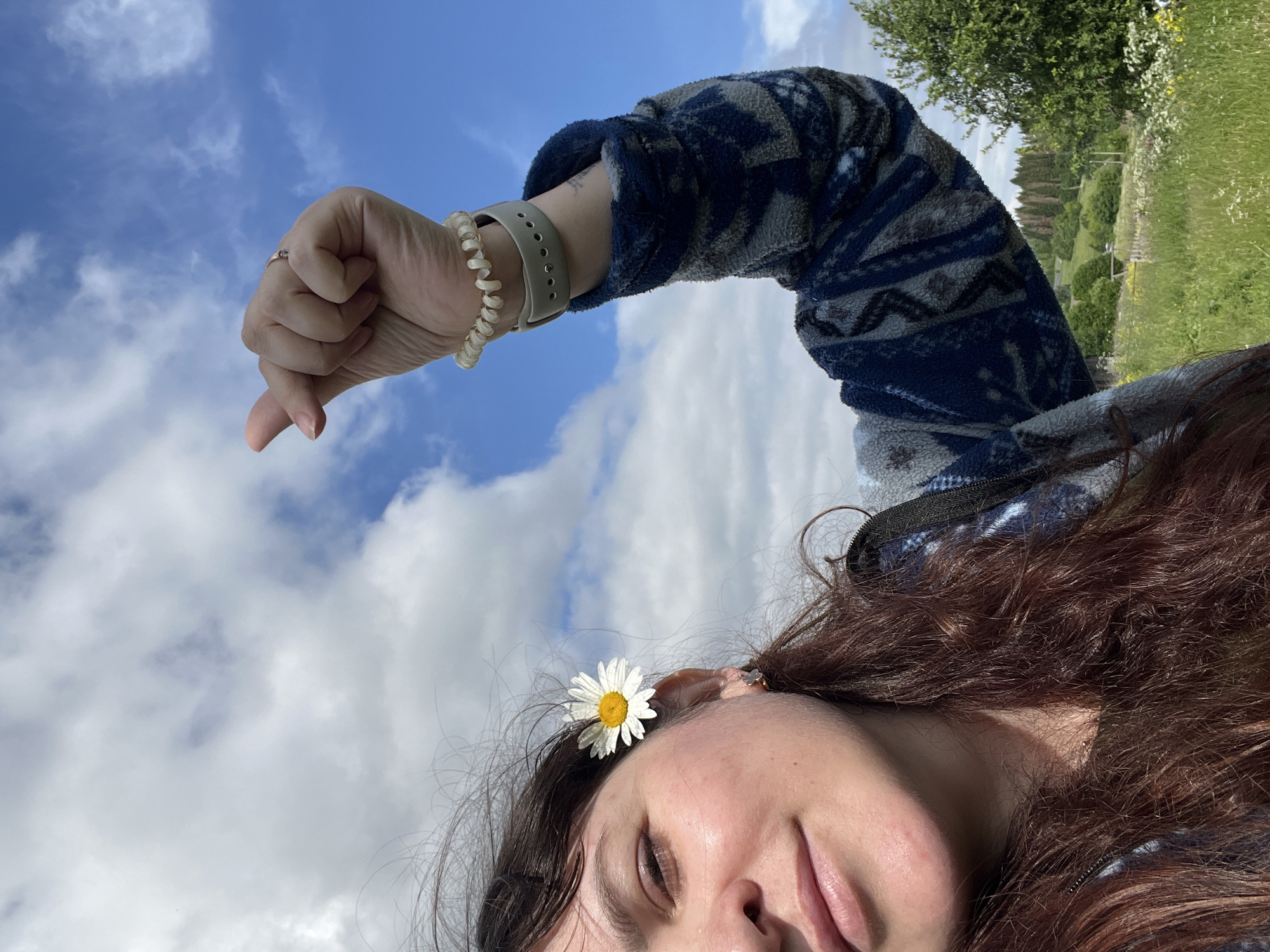 A young woman with dark, long, wavy hair is captured in a selfie outdoors. A white daisy with a yellow center is tucked behind her left ear. She is wearing a dark blue and grey patterned fleece jacket. Her right arm is raised, and her hand is making a gesture with her thumb and index finger touching, forming a heart shape. She is also wearing a thin, gold ring on her right ring finger and a beige spiral bracelet and a beige watch on her right wrist. The background shows a bright blue sky with scattered white clouds, a green grassy field with some wildflowers, and a line of trees in the distance. The location is the city of Panteleevka, Russia.