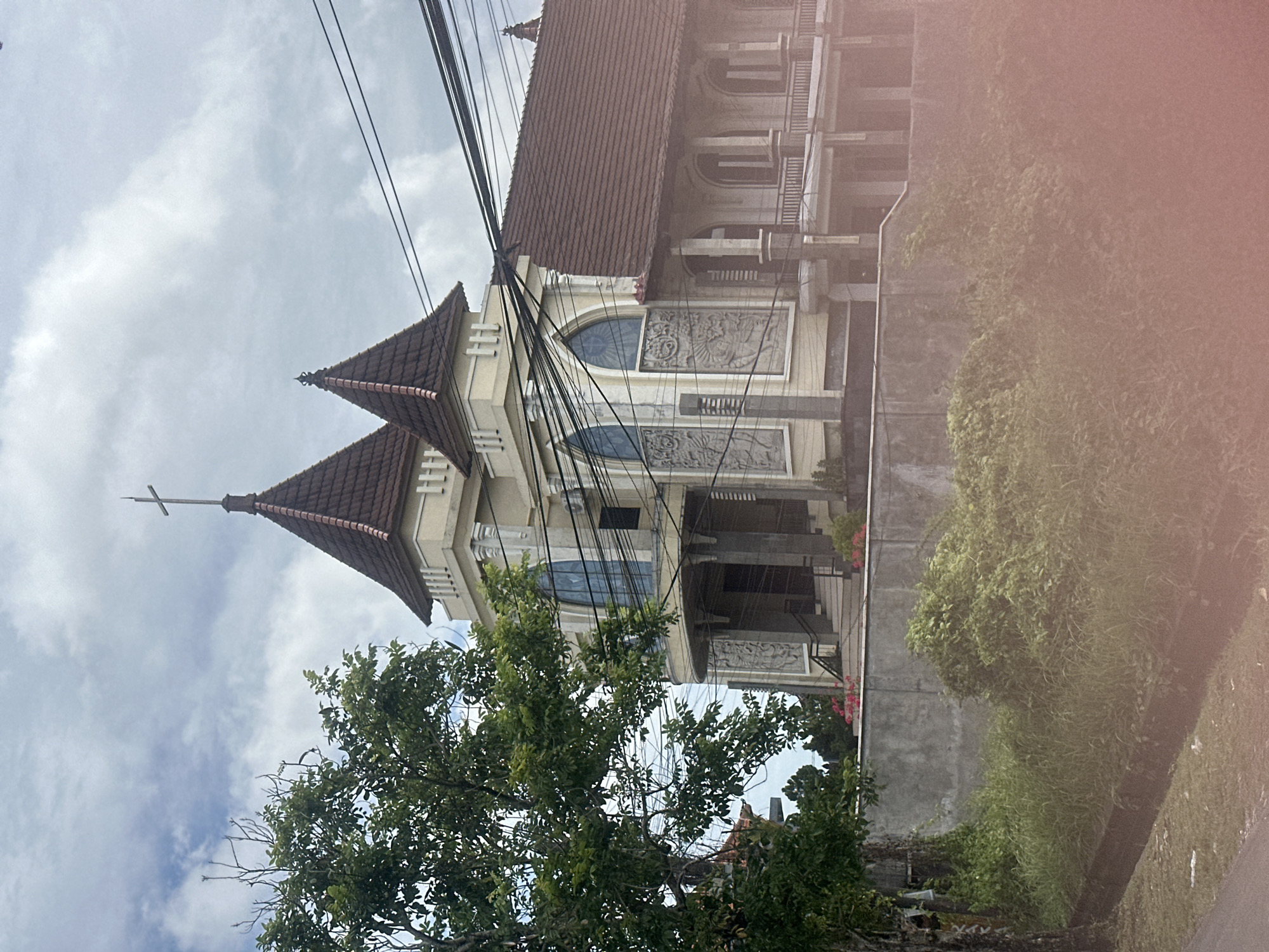 This image depicts a church with a distinctive architectural style located in North Kuta, Indonesia. The church features two pointed towers, one taller than the other, both topped with crosses. The building is painted a light yellow and has a reddish-brown tiled roof. Elaborate relief carvings adorn the exterior walls beneath arched windows, which appear to be stained glass. A balcony with decorative railings runs along the upper level of a section of the building. Electrical wires are prominently strung across the upper right portion of the frame, obscuring some of the sky.
In the foreground, a large tree with green foliage is situated to the left of the church. A concrete wall separates the church grounds from a lower area overgrown with lush green grass and bushes. A narrow, dark ditch or drain runs along the edge of the paved area in the bottom left of the image. The sky is overcast with scattered clouds. The scene appears to be captured during the daytime, possibly on a cloudy or overcast day. There are no people visible in the image.