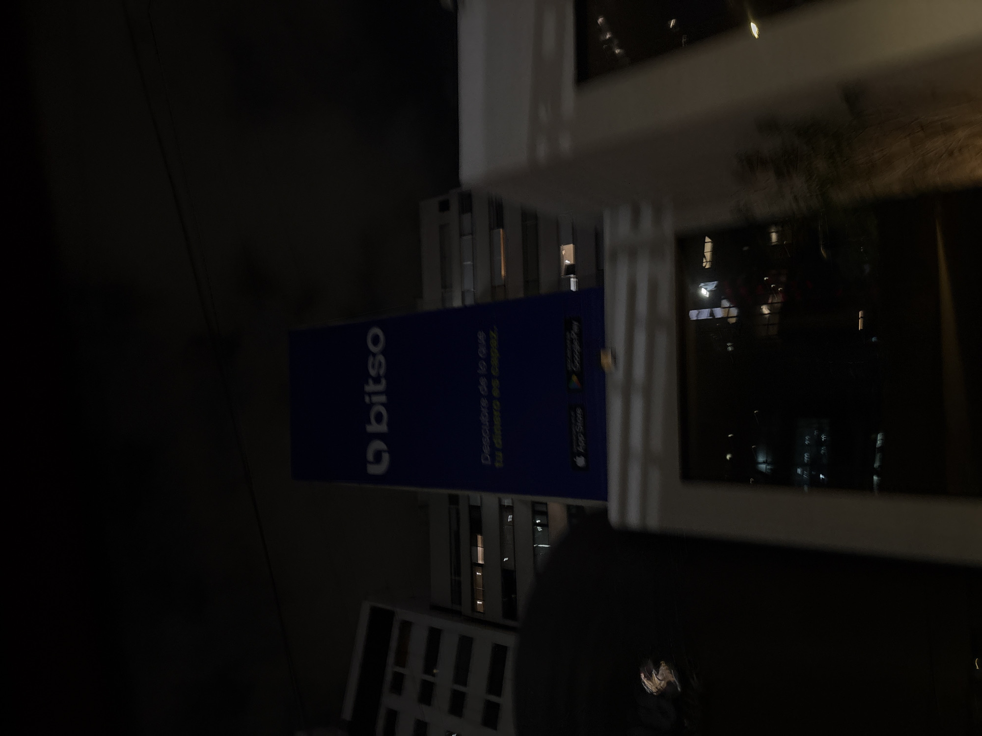 The image depicts an outdoor scene at night, likely in Mexico City, Mexico. A Bitso advertisement banner is prominently displayed, positioned in front of a building. A nearby building with lit windows is also visible in the background. A window is visible in the right of the image, providing a view to the exterior and reflecting the city lights. The lighting conditions suggest that the scene is illuminated by streetlights or other ambient sources. The composition of the image is taken from a slightly off-angle perspective, capturing the banner, the building, and a part of an office or dwelling.