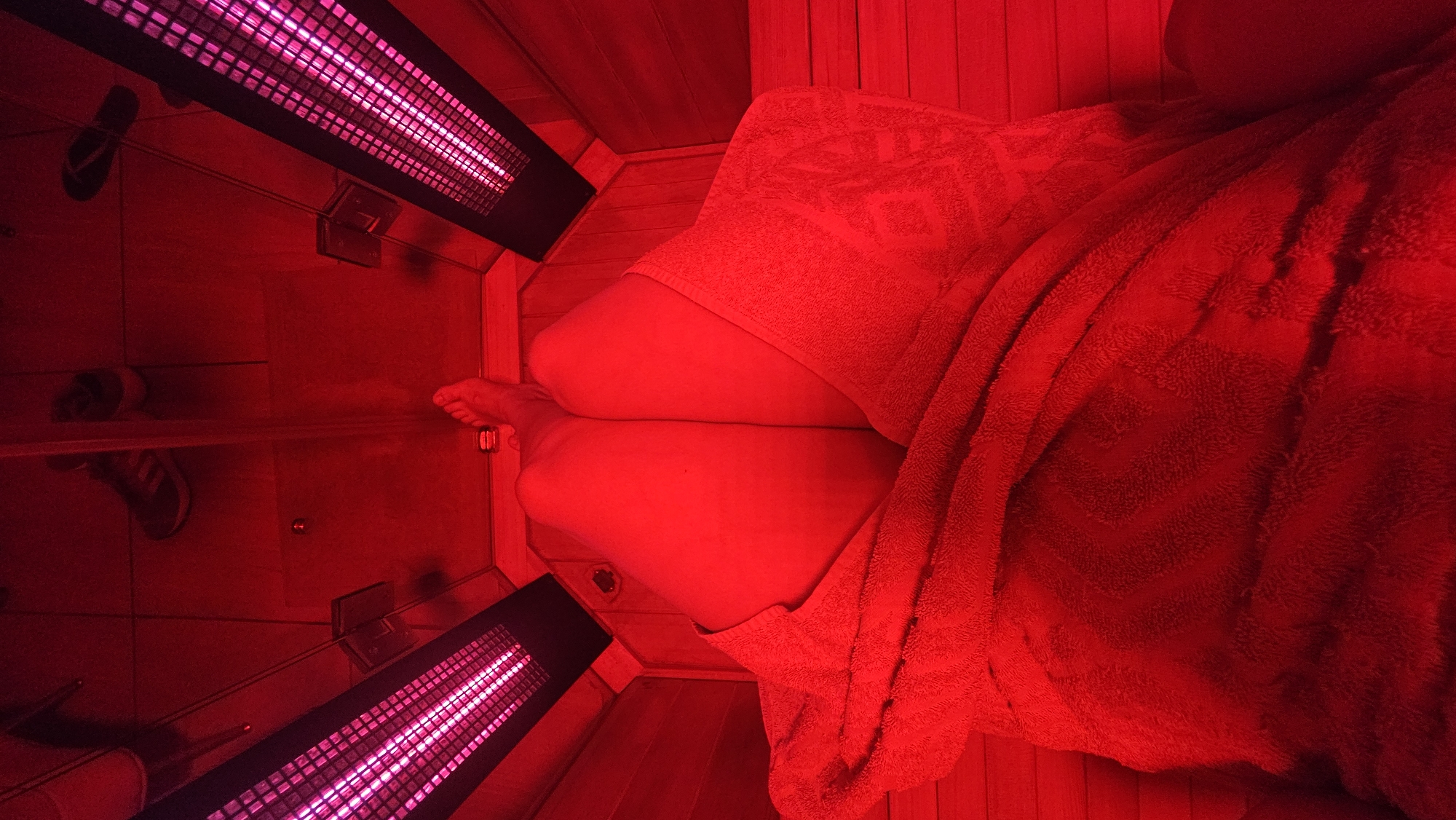 The image depicts a person inside an infrared sauna or red light therapy booth, viewed from a high angle. The entire scene is bathed in an intense red light, emanating from two long, rectangular infrared heaters positioned on the walls, one in the upper left and another in the lower left of the frame. These heaters glow with a bright pinkish-purple light.
The subject is a person, likely female, whose legs are bent, and feet are visible towards the center. Their upper body and lap are covered by a red towel or blanket, draped loosely. The walls and possibly the floor of the enclosure appear to be made of wood, though its natural color is obscured by the strong red illumination.
The activity taking place is an infrared sauna or red light therapy session, as indicated by the specific heaters and the enclosed setup. The posture of the person suggests relaxation.
Notable details include the overwhelming red light coloring everything, the visible grid pattern of the heating elements, and a small circular vent or drain on the floor. On the left side of the image, appearing outside the immediate enclosure or reflected on a surface, are a pair of black flip-flops and a black and white sneaker. No text is visible. The location context is Puebla, Mexico.