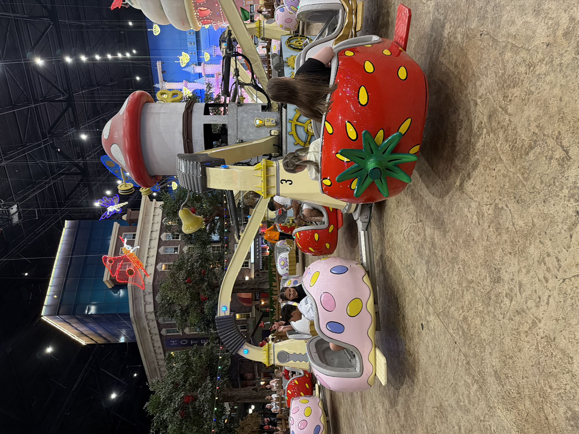 An indoor amusement park scene features a spinning ride with several passenger pods designed as oversized fruit. In the foreground, a red strawberry-shaped pod carries two young individuals. To the left, a pink polka-dotted pod with yellow and blue circles holds a child. In the background, a structure resembling a mushroom tower with white spots on a red cap is part of the ride's central mechanism. The ride is situated on a textured floor resembling stone or concrete. Above, the ceiling of the large indoor space is visible, with industrial lighting fixtures and support beams. Decorations, including trees and buildings designed to resemble a street scene, are present in the midground, along with illuminated ornamental butterflies and other festive elements. Several other people are visible around and on the ride, some appearing to be waiting for their turn. The overall environment suggests a family entertainment center or indoor theme park.