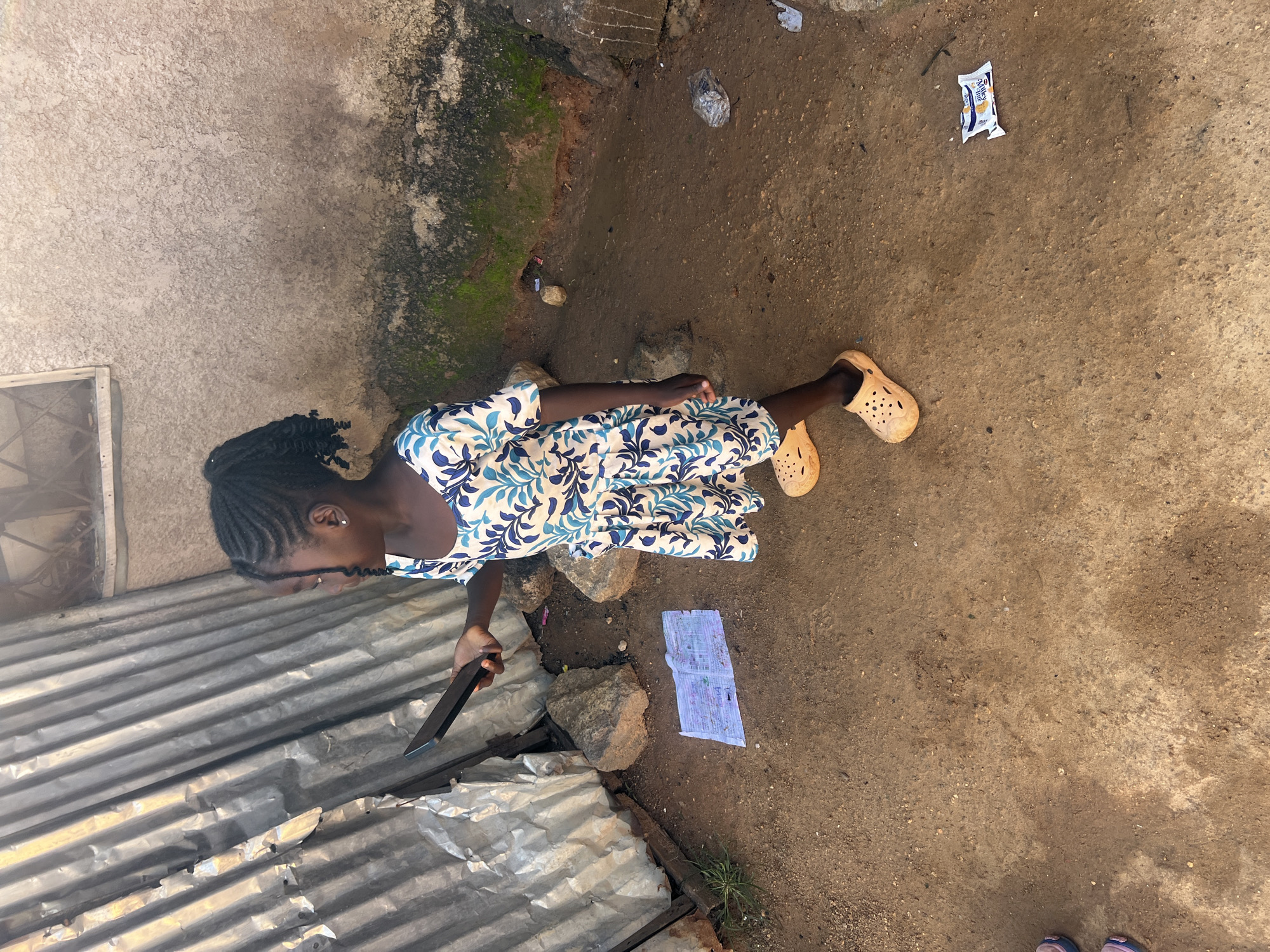 A young Black girl with braided hair, wearing a blue and white floral print dress and orange slip-on shoes, stands on a dirt path. She is holding a dark-colored object, possibly a phone, in her right hand. A crumpled piece of paper, appearing to be a notebook or document, lies on the ground in front of her. To her left is a corrugated metal structure, and to her right is a textured, light-colored wall. The ground is covered in dirt and small rocks, with some green vegetation growing near the wall. The scene appears to be an outdoor environment in a residential area, likely in the city of Jos, Nigeria, characterized by informal structures and natural ground cover.