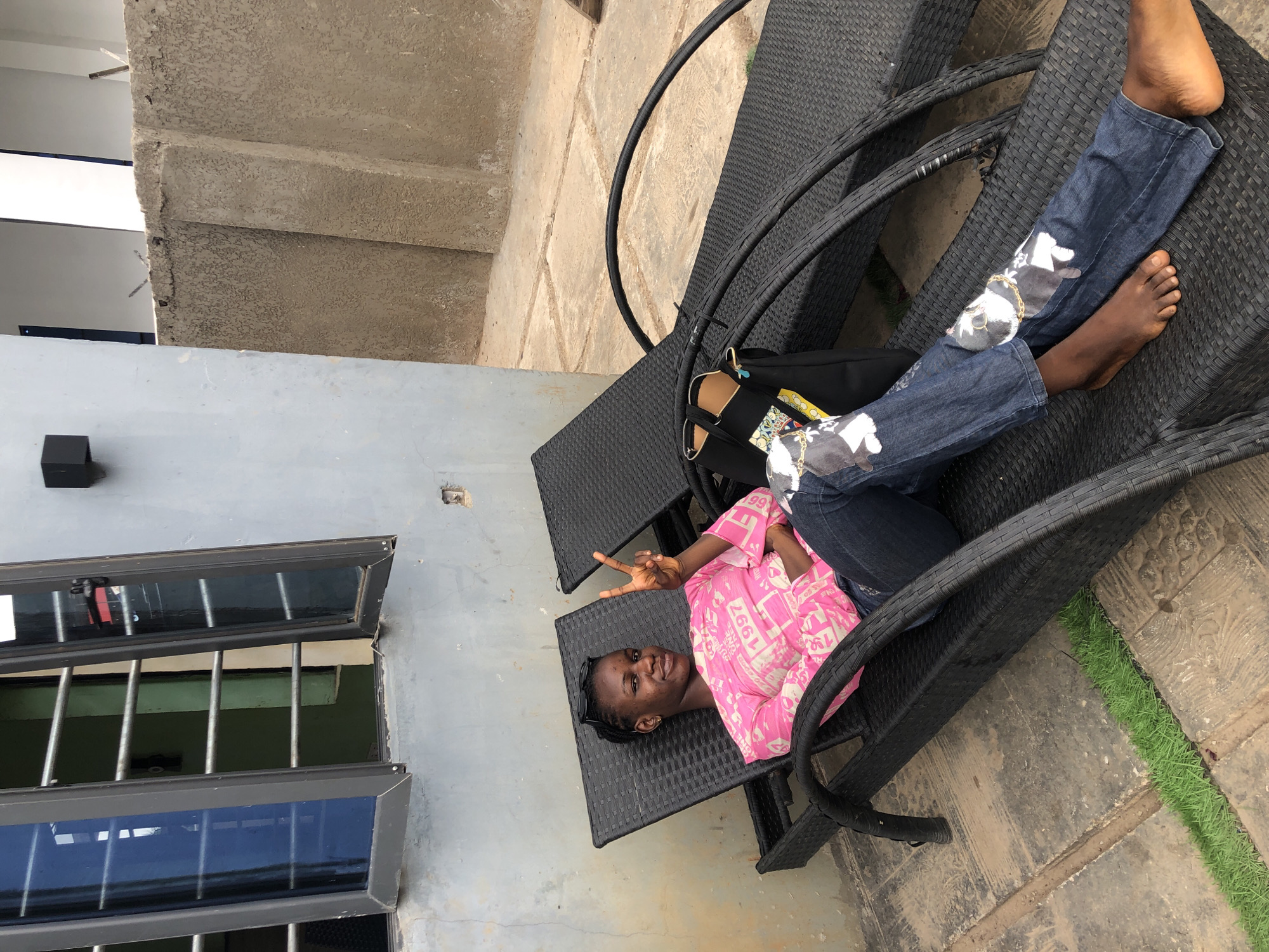 A person is lying on a black wicker chaise lounge outdoors in Akwanga, Nigeria. They are wearing blue jeans with a design and a pink and white patterned shirt. The person is barefoot, and is holding up a peace sign with their left hand. A black bag is next to them on the chaise lounge. In the background, there is a light gray building with an open window featuring a security bars. A concrete structure is visible in the upper left corner of the image. Artificial grass is also visible near the chaise lounge.
