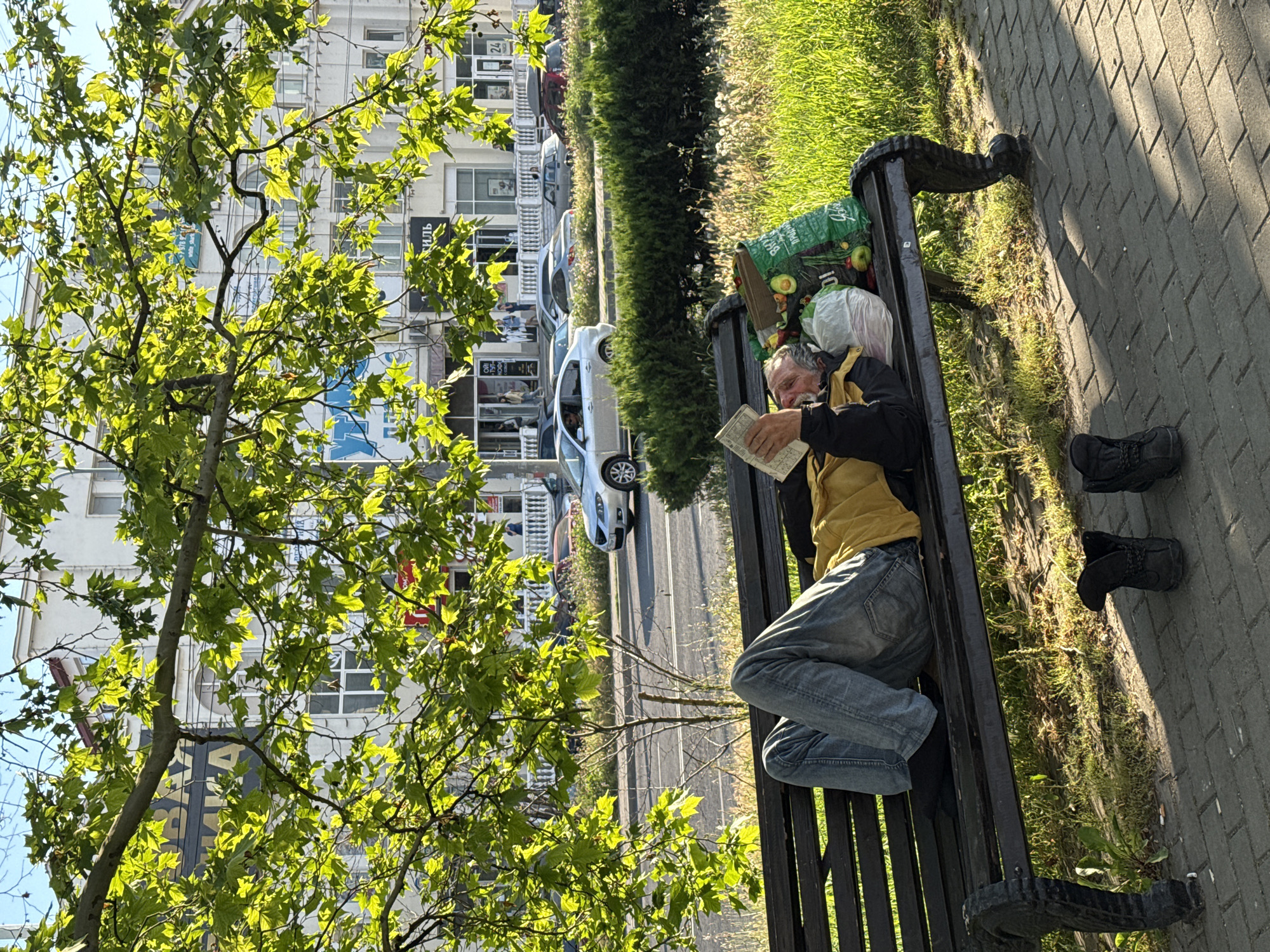An elderly man is lying on his back on a park bench, reading a book. He is wearing a yellow shirt and blue jeans. Beside him on the bench are two plastic bags filled with groceries, including apples and avocados. His shoes are placed on the ground in front of the bench. The bench is situated on a sidewalk next to a road with cars driving by and a building in the background. The scene is bathed in the warm light of the setting sun, casting long shadows.