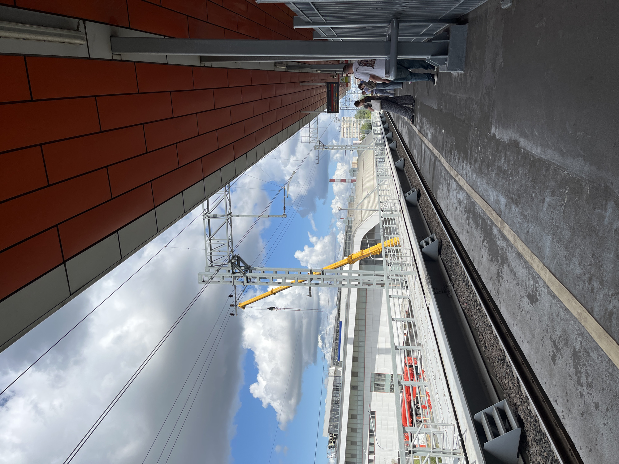 The image captures a view of a train station platform in Moscow, Russia, on a day with a partly cloudy sky. The platform is paved with concrete and bordered by railway tracks. Above the platform, an orange and white canopy extends, supported by grey pillars. Several individuals are standing on the platform, appearing to wait for a train. In the background, a modern building complex with a white facade and large windows is visible, alongside a yellow construction crane and overhead power lines for the trains. The general atmosphere suggests an active, urban transportation hub.