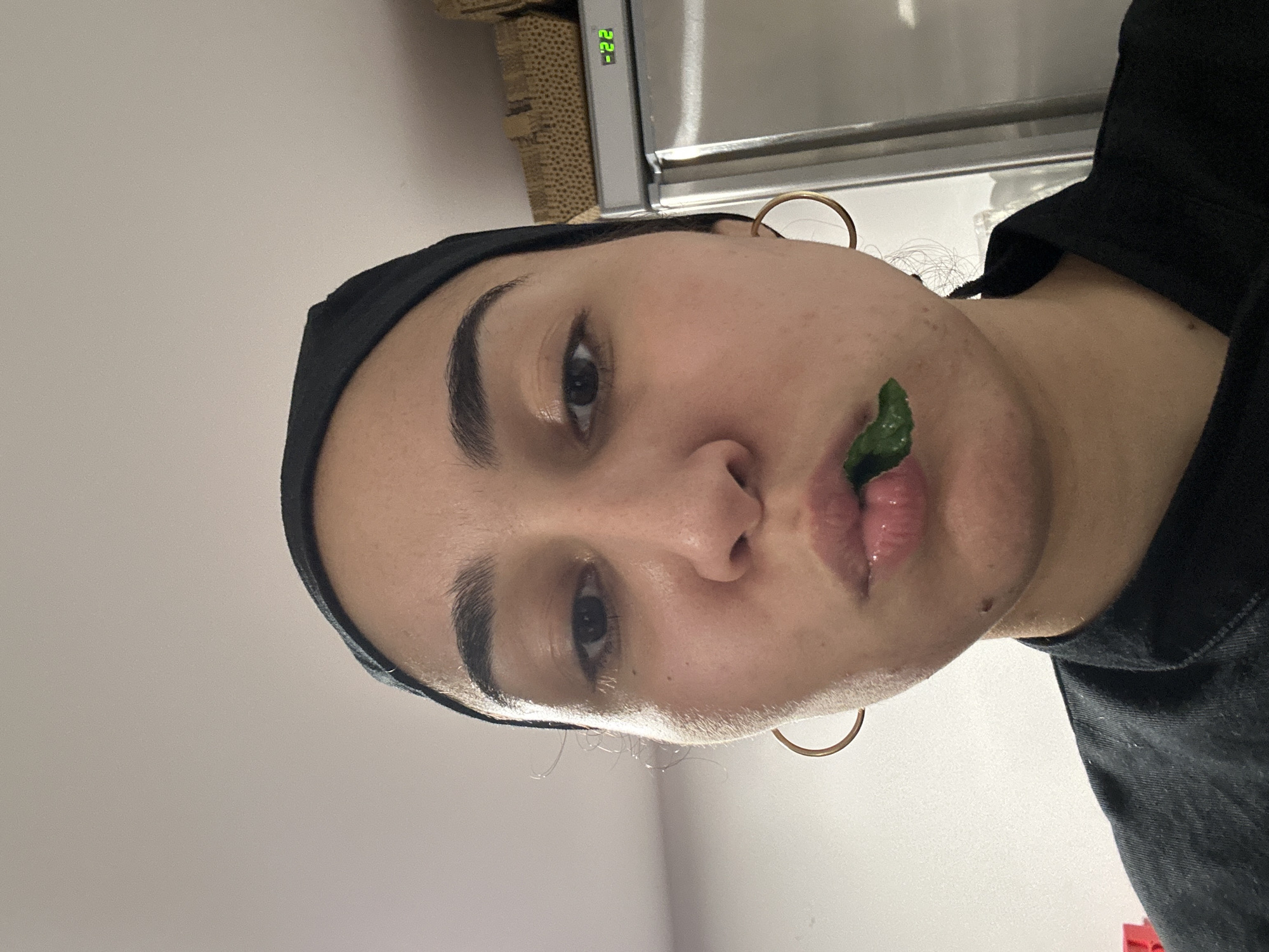 A close-up, slightly off-center portrait shot shows a person with light brown skin. They have dark eyebrows, dark eyes, and are wearing a black headband. Gold hoop earrings are visible. A small, dark green item is held in their lips. The person is wearing a dark-colored shirt or jacket. The background is a plain, light-colored wall and a stainless steel or light grey appliance. A small digital display showing "22" is visible on the appliance. No information suggests that this image was taken in Burgas, Bulgaria.