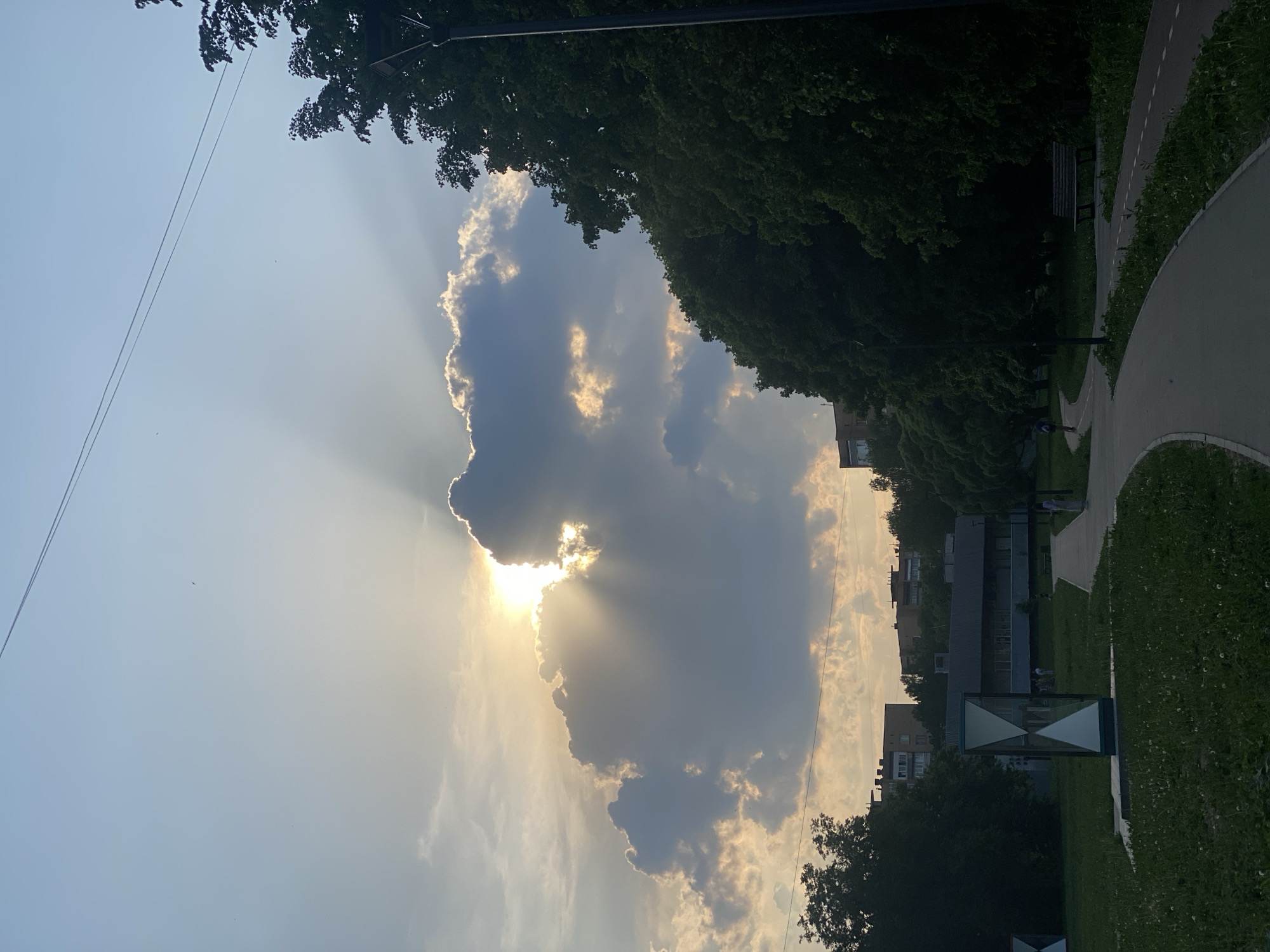 The image depicts a sun obscured by a large cloud, with sun rays radiating outwards. Adjacent to the sun and cloud is a row of trees. In the background are buildings. In the foreground is a paved pathway and greenery. There is also a sign with an hourglass shape on it. The location is in a city of Unknown, Unknown.