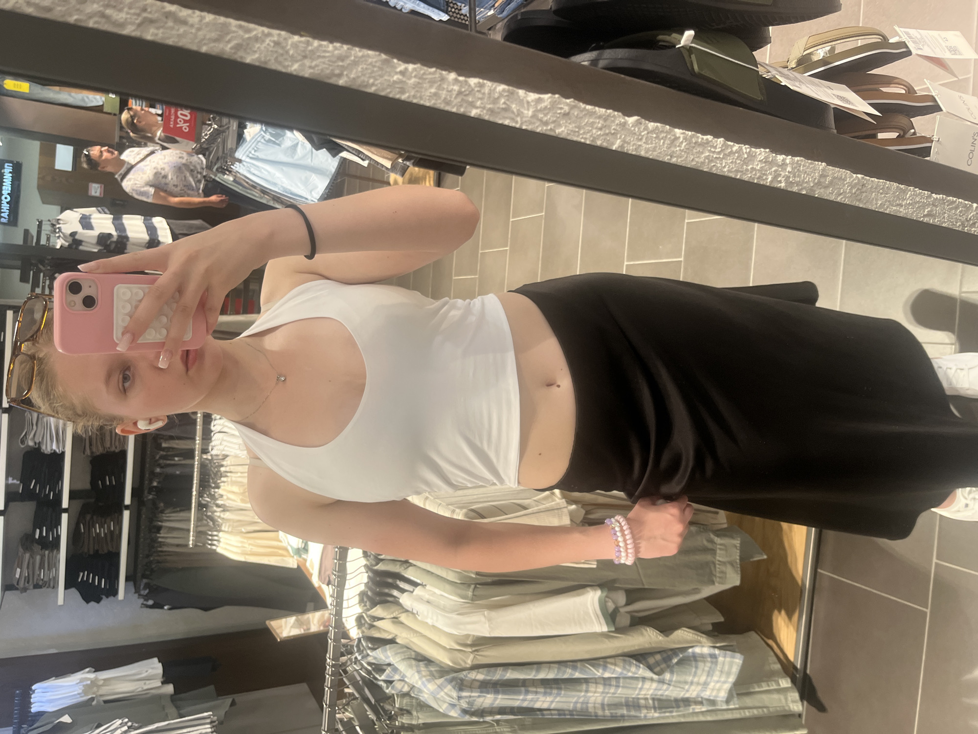 The image depicts a young woman with blonde hair taking a mirror selfie in a clothing store. She is wearing a white halter top, a black midi skirt, and white sneakers. She has a pink phone case in her right hand and is wearing white earbuds. Several racks of clothing are visible in the background, along with a few other shoppers reflected in the mirror. The store appears to be a chain clothing retailer, possibly in a shopping mall. The scene is well-lit and the woman is the primary focus.