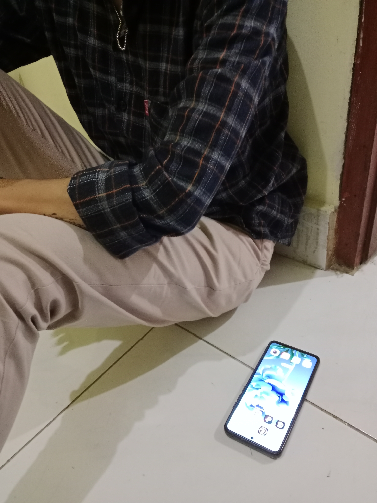 A person is sitting on the floor with their legs crossed. They are wearing a plaid shirt and light-colored pants. A smartphone is lying on the floor next to them, displaying a home screen with several app icons. The environment appears to be indoors, with tiled flooring and a wall in the background. The image does not provide any information about the location in the city of West Jakarta, Indonesia.