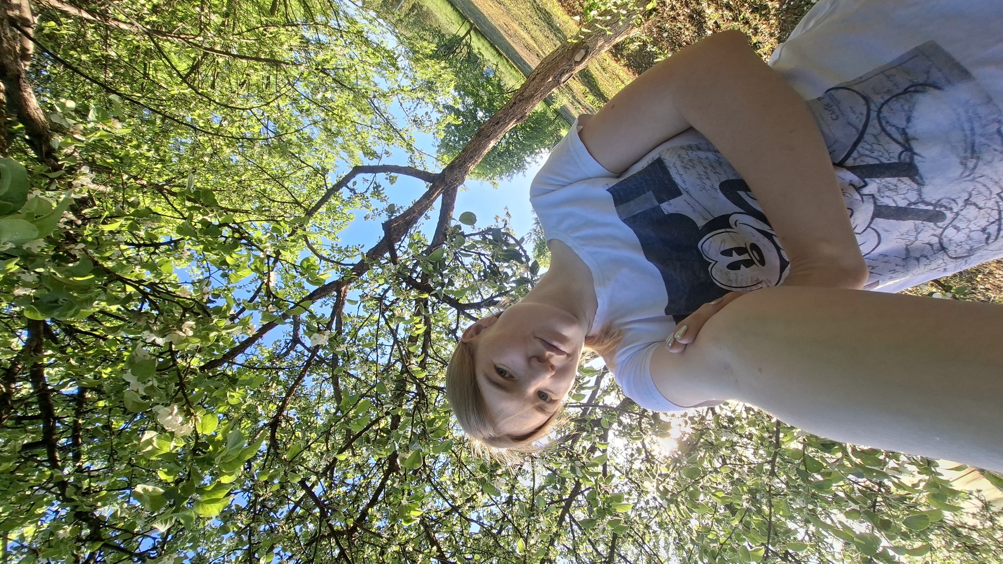The image shows a fair-skinned person in Большой Букор, Russia, lying on their back under a tree with green leaves and white blossoms. The person is wearing a white T-shirt with a Mickey Mouse graphic. The camera is positioned above the person, looking down at them and up into the tree canopy. Patches of blue sky are visible through the leaves. In the background, a field is partially visible beyond the tree.