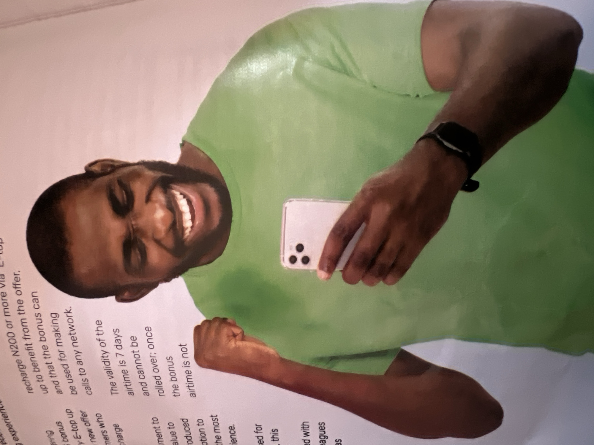 The image shows a Black man in a lime green t-shirt holding a smartphone and raising his left fist. He is laughing with his eyes closed. He is wearing a black smartwatch on his left wrist. In the background, there is text on a white surface that appears to be a brochure or flyer. The text discusses a bonus offer for recharging N200 or more via E-top up, stating the bonus can be used for calls to any network and is valid for 7 days, not rolling over. The man is positioned in the foreground, with the text to his left. The lighting is even, suggesting an indoor setting. The image quality is somewhat grainy, possibly from a photograph of a printed document. There is no identifiable information within the image that places the scene in Jalingo, Nigeria, or any specific urban location.