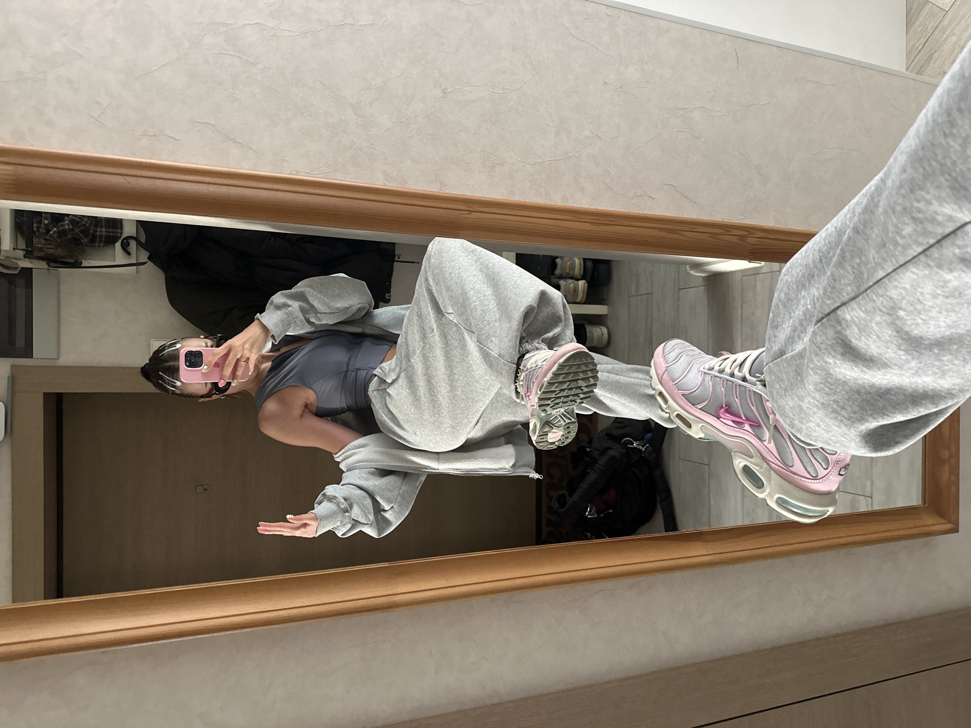 A person is taking a full-body mirror selfie in what appears to be a home or apartment hallway. The person is wearing a grey athletic set consisting of a tank top, zip-up hoodie, and baggy sweatpants. They are also wearing pink and grey sneakers. The person is holding a pink smartphone in their right hand and has their left hand raised. The mirror reflects the person and the hallway behind them, which includes a wooden door, a light-colored wall with a textured finish, and flooring that looks like light-colored tiles. In the reflection, a rack with several jackets and bags can be seen. There are no visual cues in the image to suggest this location is in Moscow, Russia.