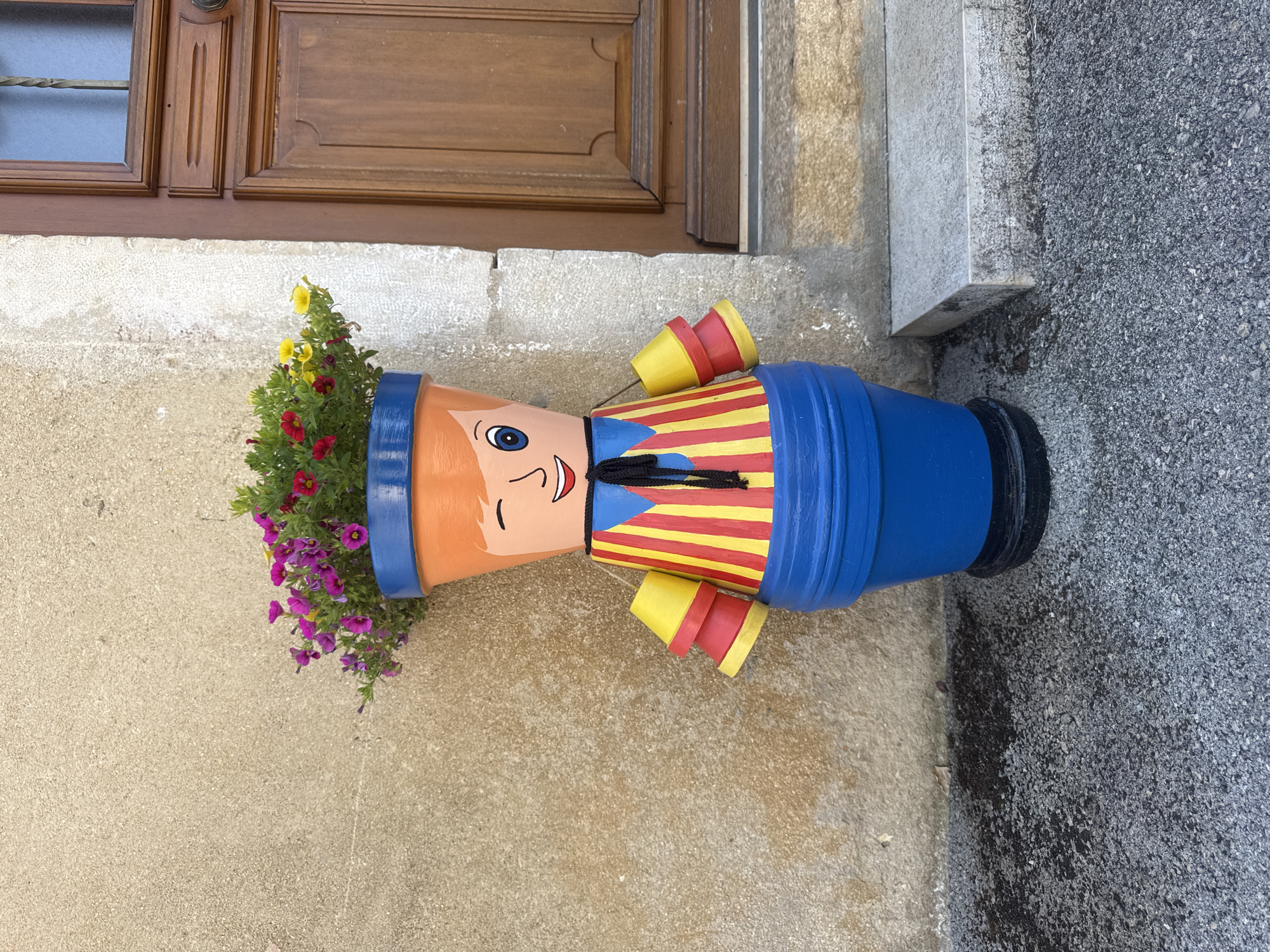 An outdoor, eye-level, medium shot captures a whimsical flower pot decoration in Coteaux du Lizon, France. The decoration, resembling a cartoonish character, is crafted from painted terracotta pots stacked and adorned with colorful details. The character's head is an orange pot with painted orange hair, a winking eye, a closed eye, and a smiling mouth. A blue rim encircles the top, holding a vibrant bouquet of pink, red, and yellow flowers. The character's torso is an orange pot painted with a blue collar and a red and yellow striped shirt with a black tie. Two yellow and red cone-shaped objects hang from strings on either side of the torso, mimicking arms. The base of the decoration is a large blue pot. The character stands on a dark circular base on a gravelly, dark grey ground. Behind the decoration, a textured, light beige wall is visible, with a portion of a dark brown wooden door and a stone step to the right. The overall scene suggests a decorative garden or entryway in a French town.
