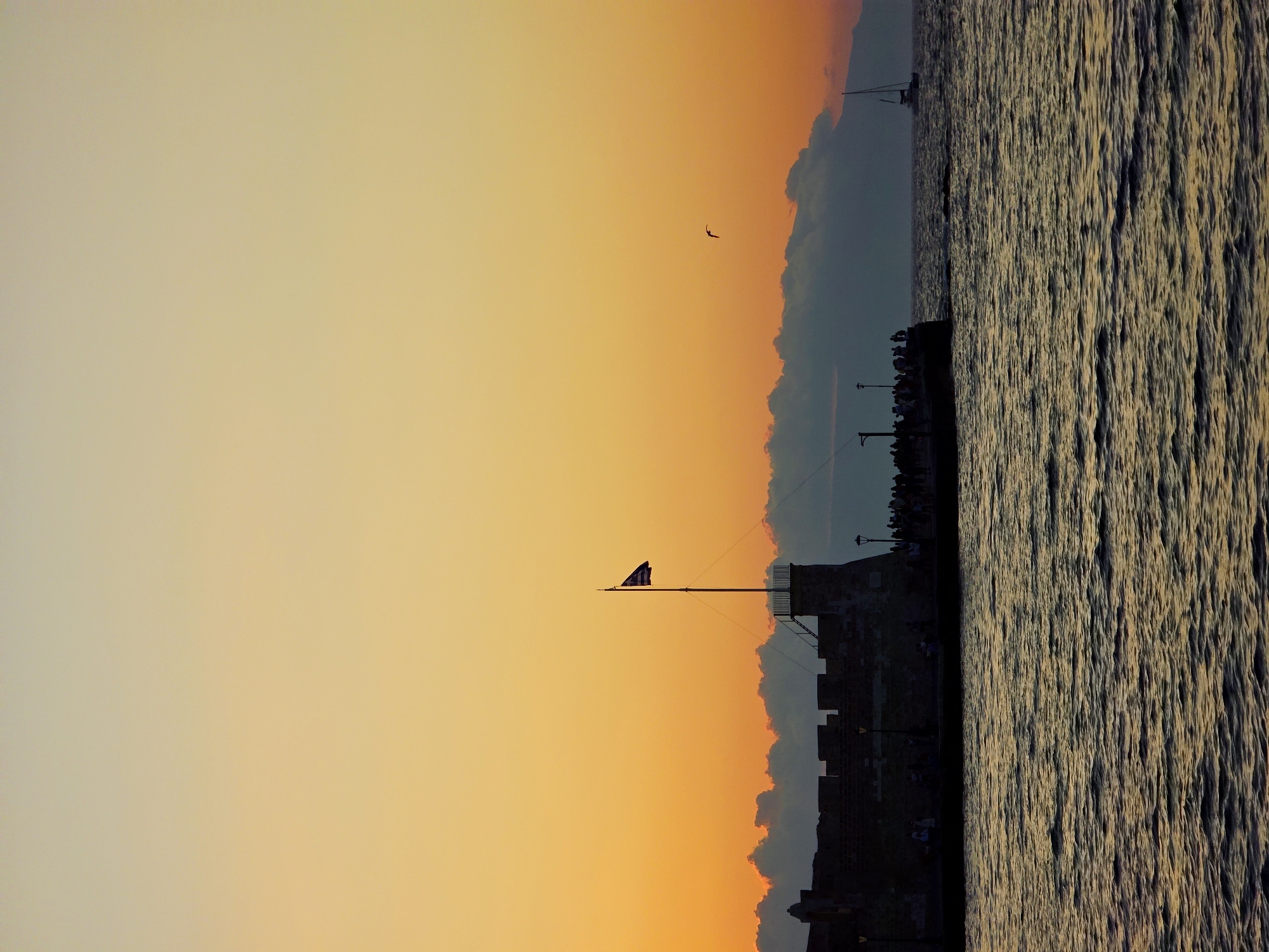 The image depicts the Venetian harbor of Chania, Greece, during a sunset. A stone fortress wall dominates the left side of the frame, with a flagpole flying the Greek flag centrally located. A group of people are silhouetted along the top of the wall, observing the sunset. The sea in the foreground is choppy, reflecting the warm hues of the sky. A sailboat is visible in the distance on the right. The sky transitions from a pale yellow at the top to a vibrant orange and pink near the horizon. The overall mood is serene and picturesque.