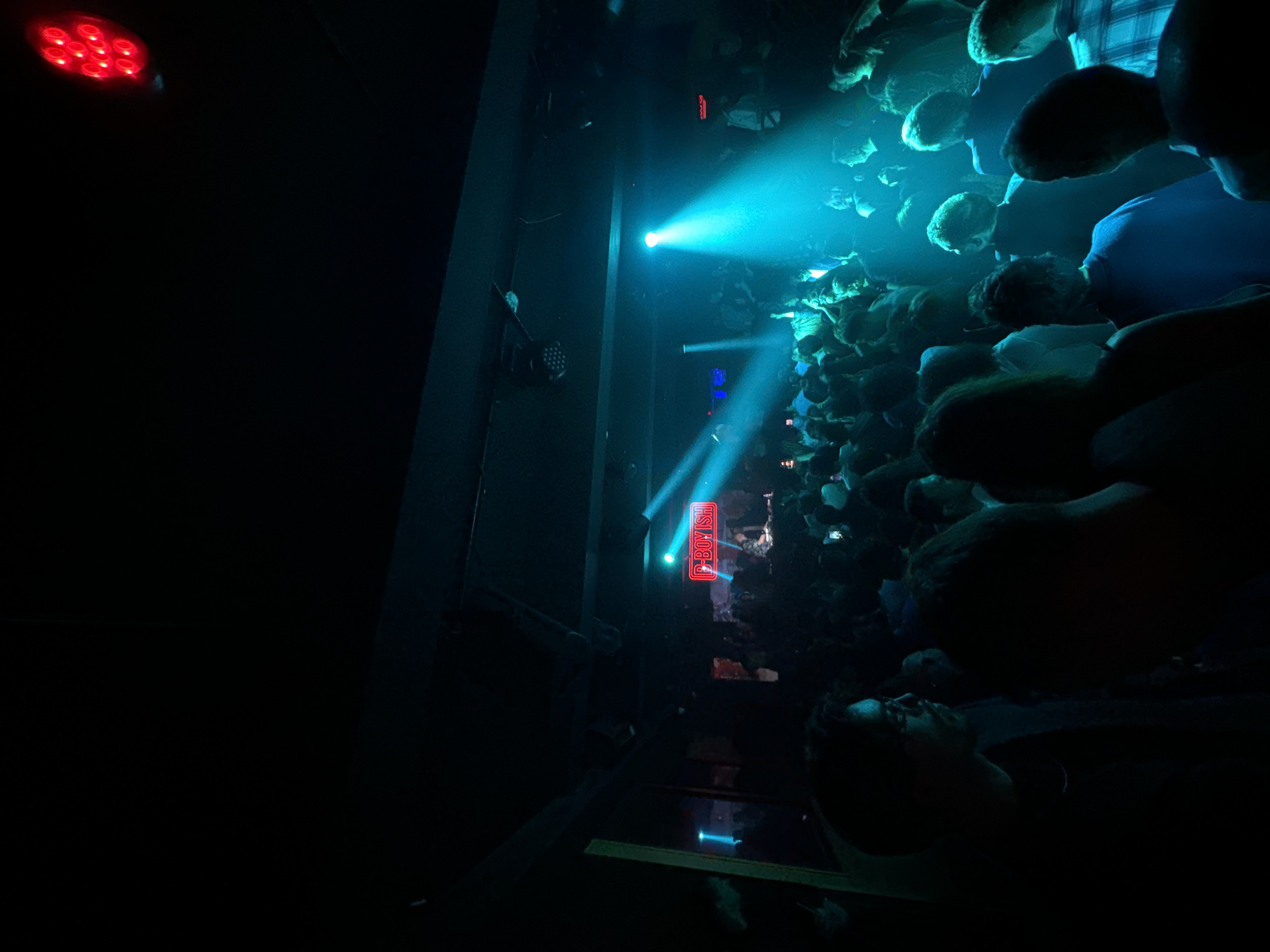 The image depicts a crowded concert venue, possibly in Paris, France. The lighting is predominantly dark with intense blue and red spotlights illuminating the scene. A large crowd of people is visible, primarily consisting of dark shapes with indistinct features, suggesting a low-light environment. A neon sign with the text "C-BOYISH" is illuminated. The venue appears to be enclosed, with dark walls visible on the left side of the frame. A red stage light can be seen in the upper left corner.