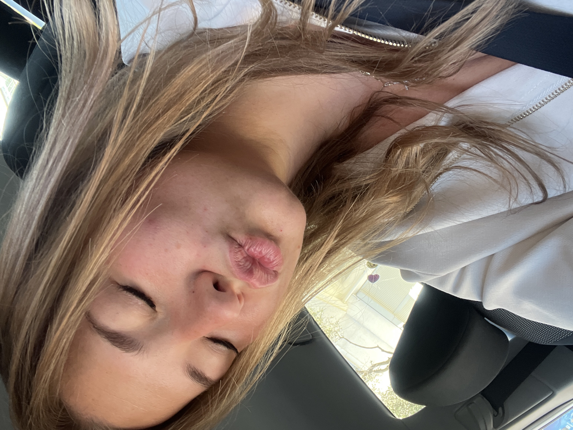A woman is inside a car, seen from a low angle. She has her eyes closed and puckered lips in a kissing expression. She has fair skin, light brown hair, and is wearing a light-colored jacket with a dark top underneath. A silver necklace is visible. The car's interior is visible, with a black headrest and a partial view of the car's window, showing trees outside. There's no information available in this image to determine that the woman is in Budva, Montenegro.