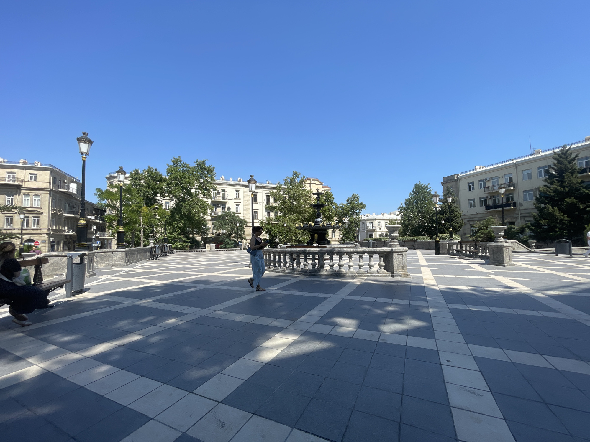 The image shows a wide shot of Fountain Square in Baku, Azerbaijan, on a clear, sunny day. The square is paved with a distinctive pattern of grey and white tiles, with geometric lines creating a visually striking effect. The area is flanked by several buildings with classic architectural styles, featuring ornate facades, balconies, and numerous windows. Trees with lush green foliage are interspersed throughout the square, providing shade and softening the urban landscape.
In the foreground, on the left side, a woman is seated on a park bench, facing away from the camera. She is dressed in dark clothing and has a light-colored bag on her lap. Further into the square, a lone woman walks across the tiled expanse, wearing a dark tank top and light blue pants, carrying a blue purse. She is walking towards a decorative fountain that is partially visible in the mid-ground. The fountain is surrounded by a stone balustrade with ornamental urns.
Several lampposts with ornate black ironwork are positioned around the square, adding to the classic aesthetic. The sky is a vibrant, cloudless blue, indicative of good weather. Shadows from the trees and possibly from overhead structures are cast across the tiled pavement, creating a dappled effect. The overall scene conveys a sense of a well-maintained public space within a historic city.
