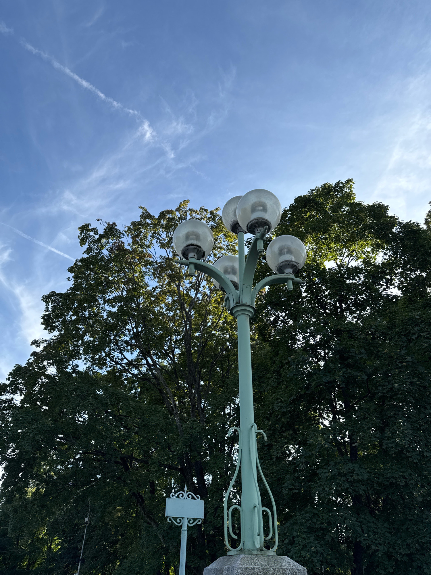 A green, ornate lamppost with five spherical globes stands in front of a dense canopy of green trees under a bright blue sky with thin white clouds and a contrail. A small, decorative white sign is visible on a pole to the left of the lamppost. This scene appears to be located in Saint Petersburg, Russia, possibly in a park or along a boulevard.
