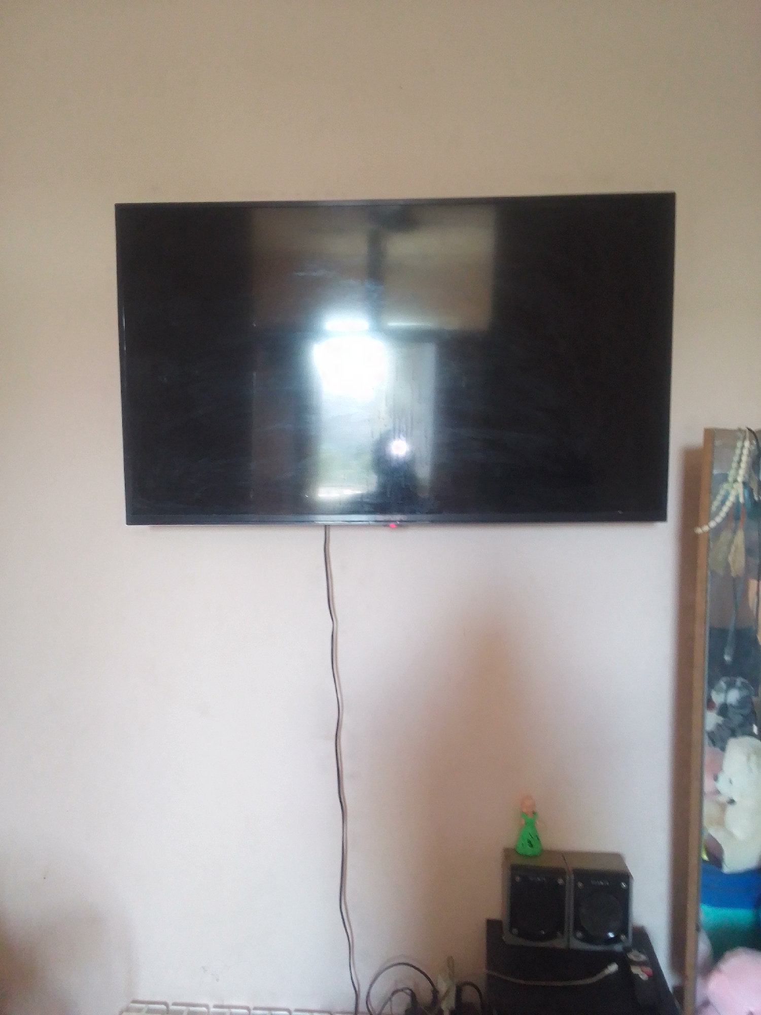 In an indoor setting, a large, flat-screen television is mounted on a pale pink wall. The screen is black, reflecting light from an unseen source. A thin black cord descends from the bottom center of the television and trails down towards the floor. To the right of the television, a portion of a wooden cabinet is visible, adorned with various items including a strand of white beads. Below the television and to the left, a section of a white radiator with a ribbed design is visible. Further to the right, on a dark surface, sit two small black speakers and a green figurine of a person. The overall environment appears to be a domestic living space. The location is the city of Kattakurgan, Uzbekistan.
